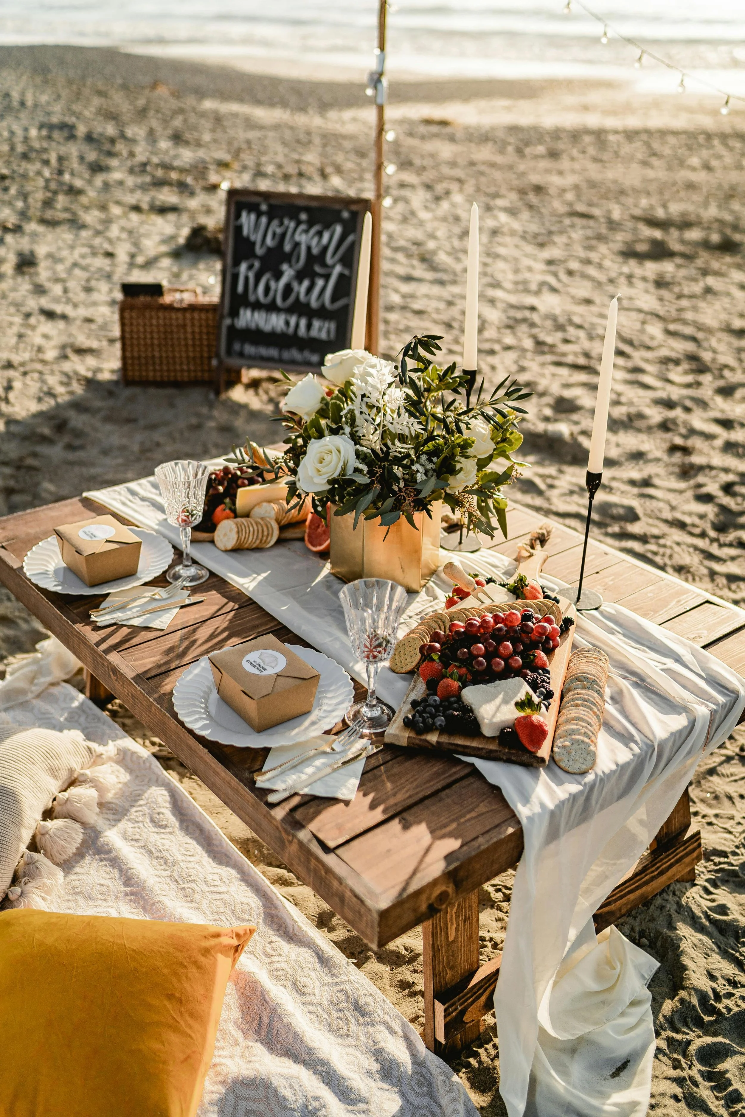 Beachside decorated table with flowers, candles, and food, set for a gathering or celebration, with a chalkboard sign in the background reading "Morgon Rout" and the date "January 8, 2023."