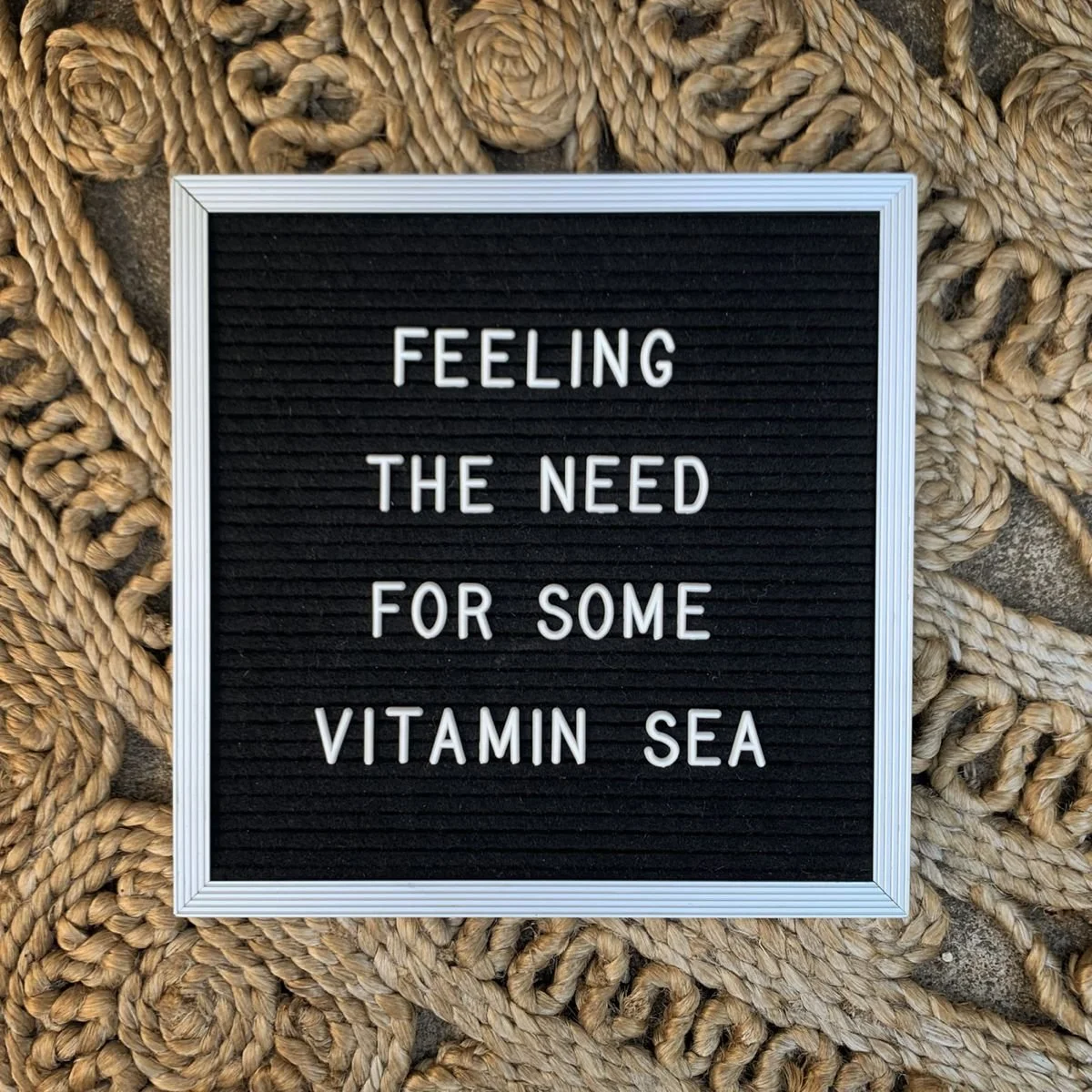 A black letter board with white text that reads, "Feeling the need for some vitamin sea," placed on a large, coiled rope.