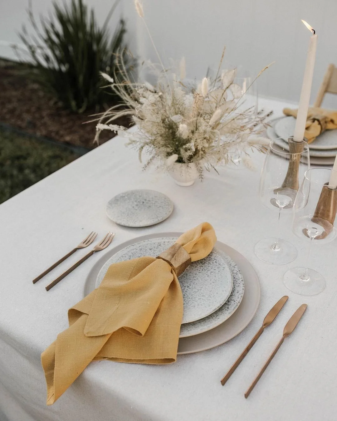 Elegant outdoor dinner table setup with white tablecloth, textured plates, yellow napkin, gold cutlery, and a floral centerpiece of dried white and beige plants with tall candles in glass holders.