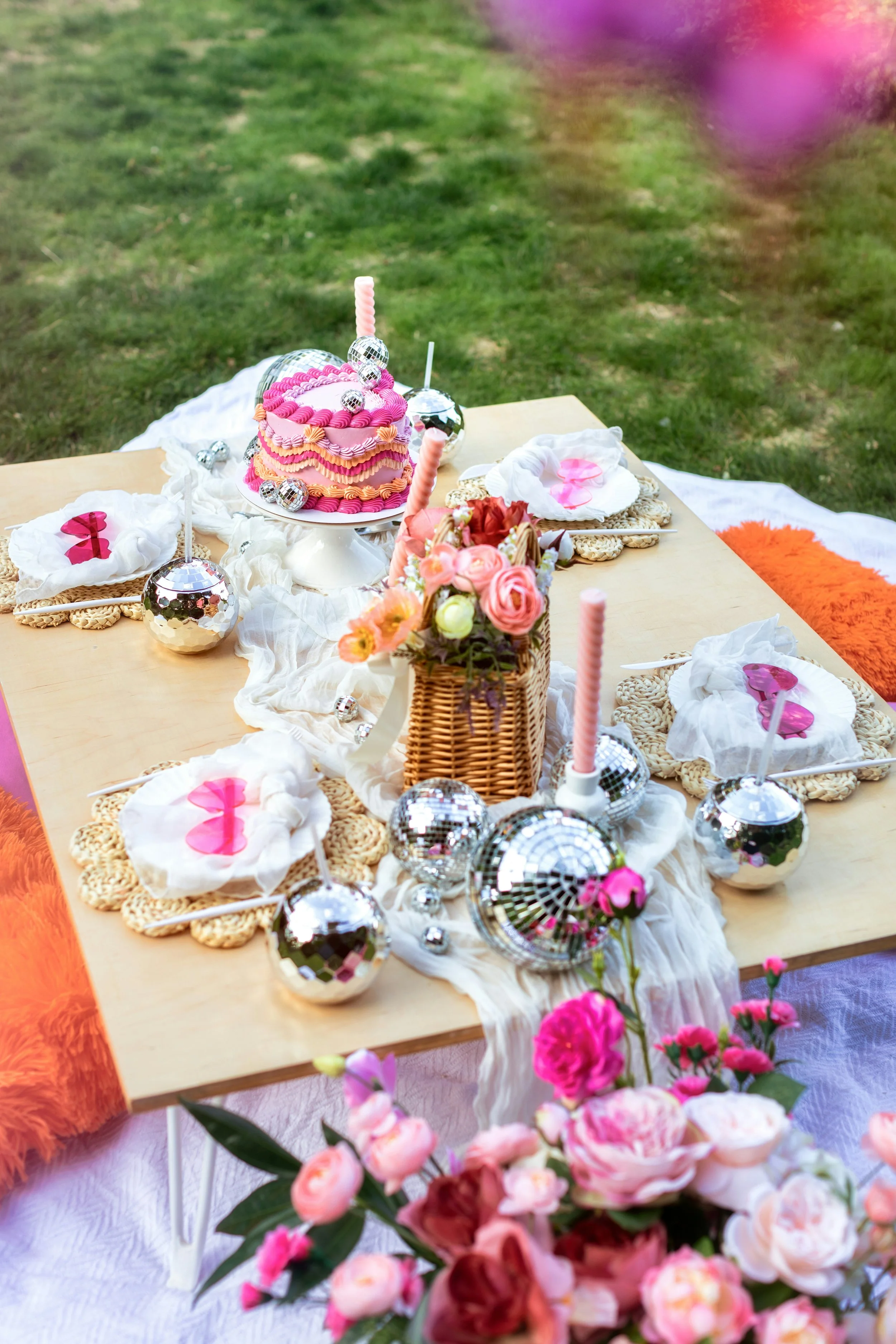 A pastel-colored outdoor birthday party table decorated with pink and white confectionery, floral arrangements, and disco ball ornaments on a grassy yard.