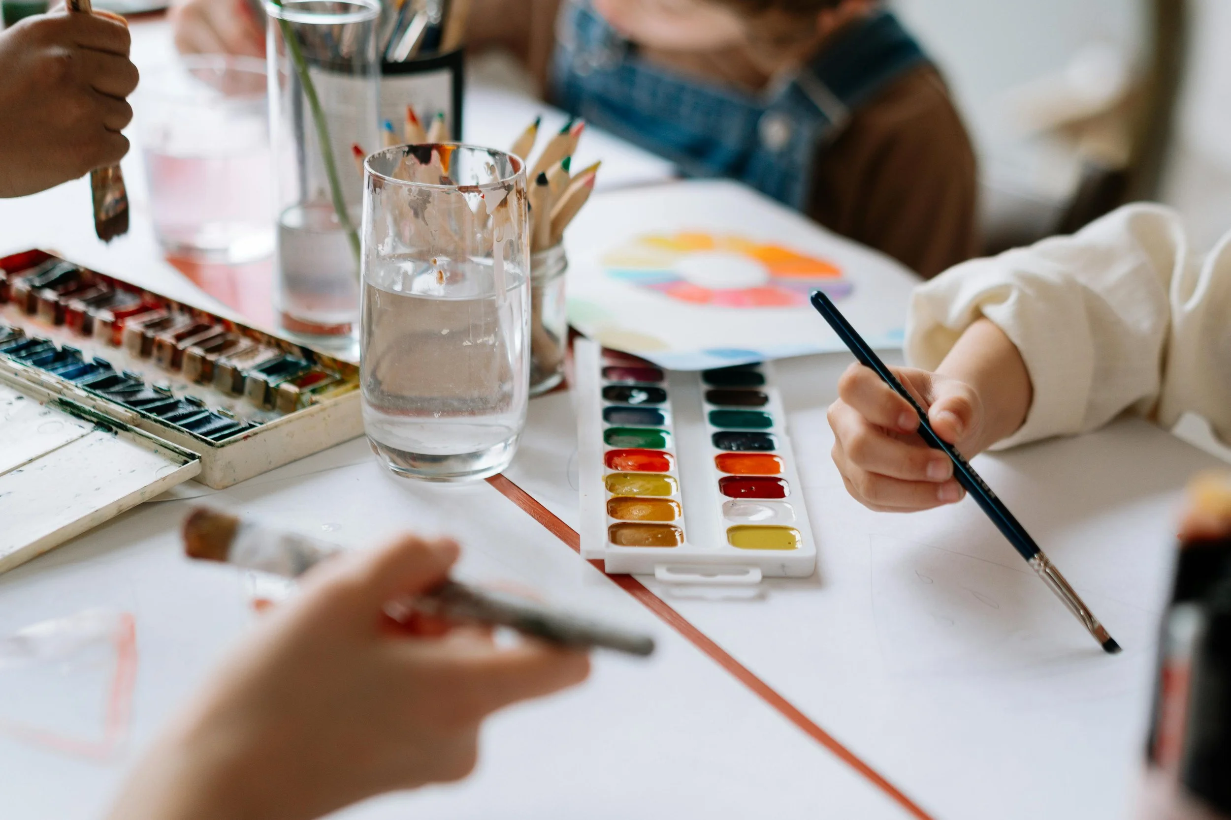 Children participating in a watercolor painting activity, with paint palettes, brushes, and a glass of water on the table.