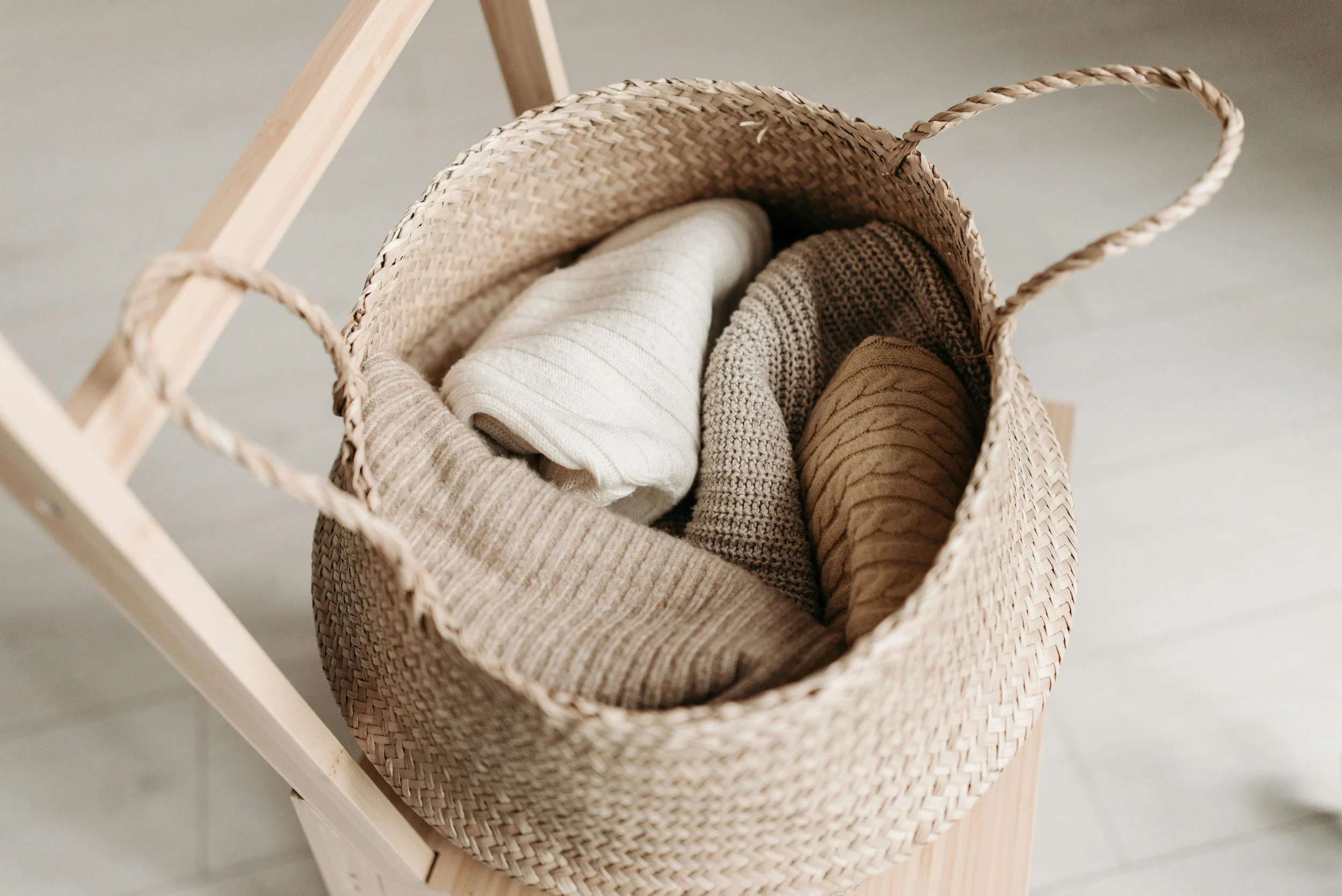 A woven straw basket containing folded cream, beige, and taupe knitted and fabric clothing items, resting on a light wooden stool.