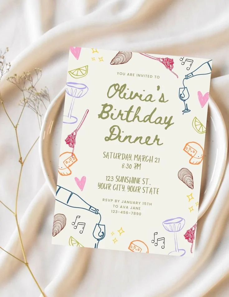 Colorful birthday dinner invitation on a white plate, surrounded by dried decorative plants. The invite features illustrations of wine glasses, lemon slices, cheese, croissants, hearts, and musical notes.