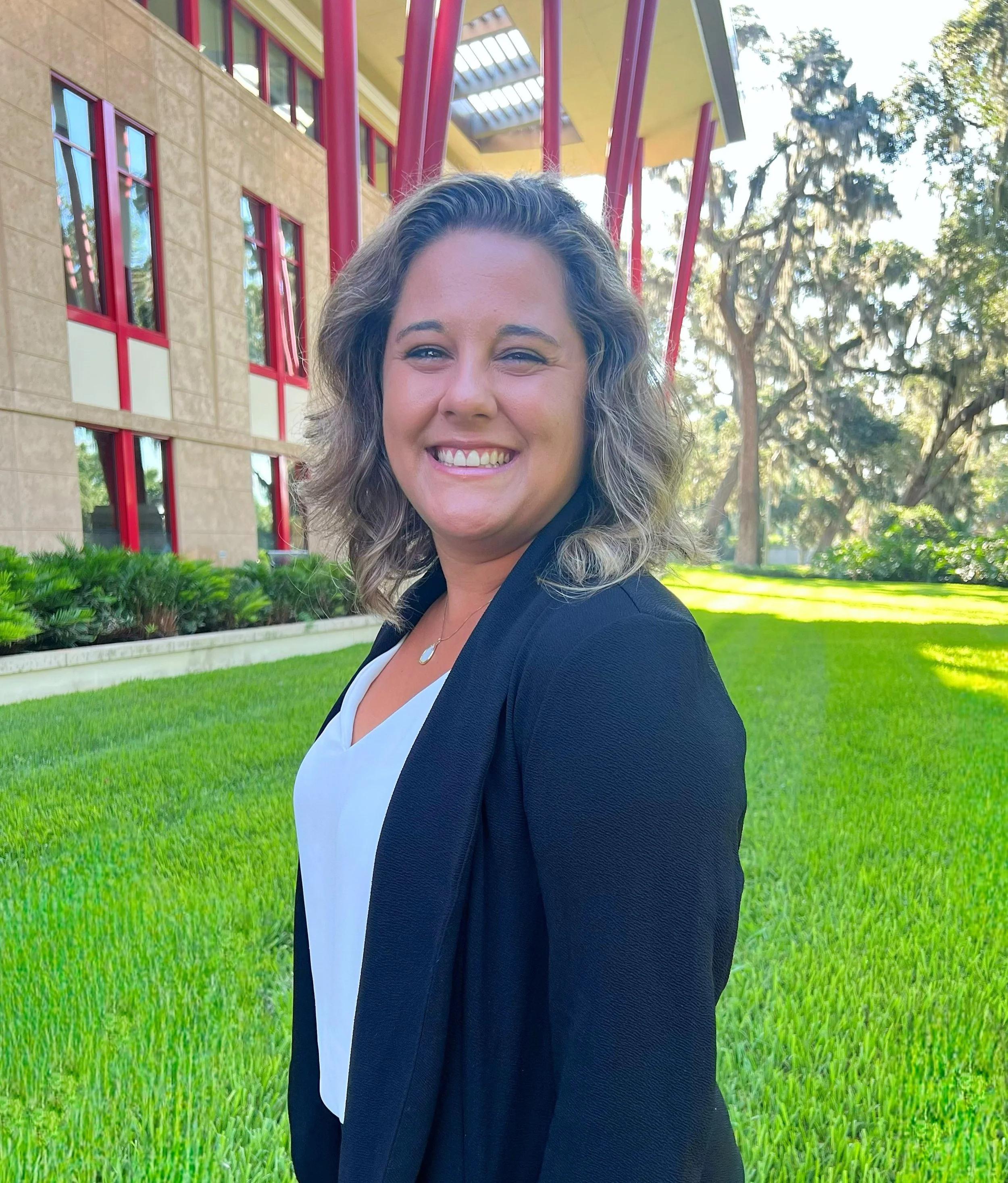 Dr. Victoria Lew, a woman smiling, with curly hair wearing a black blazer over a white blouse, standing outdoors on a sunny day with green grass and a building with large windows and red accents in the background.