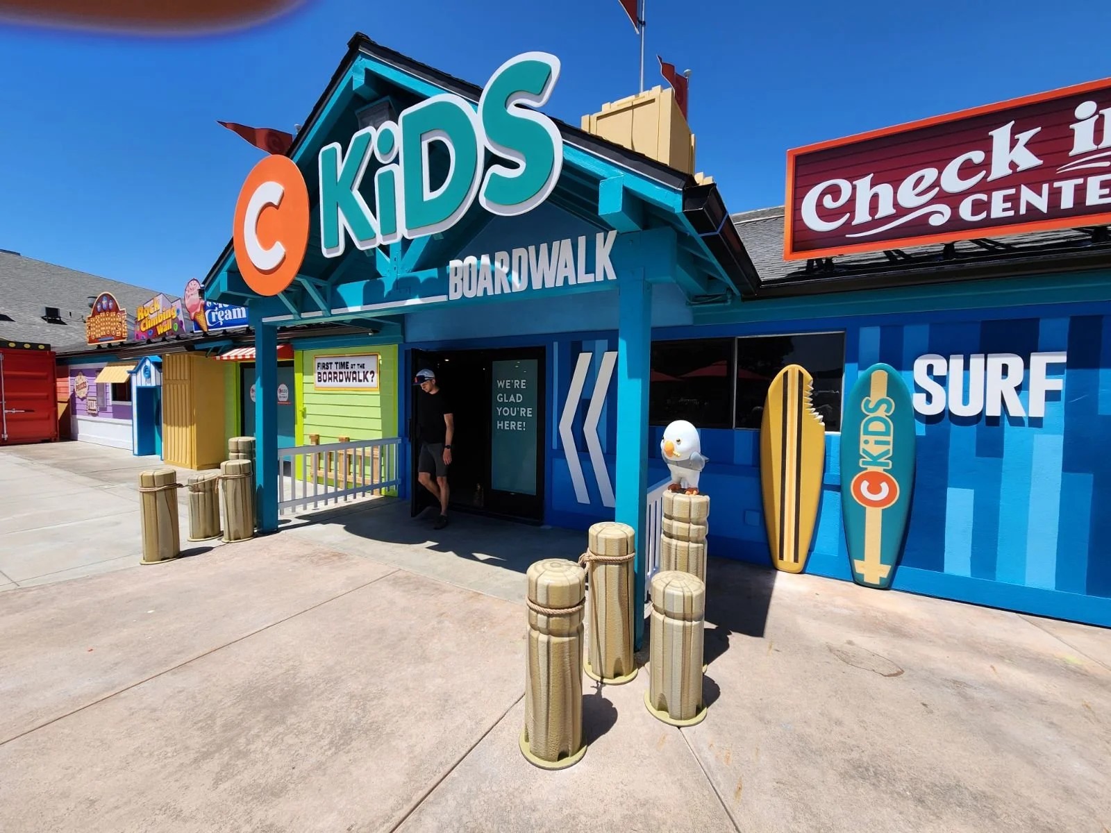 Entrance of a colorful Chuck E. Kids Boardwalk with a person walking out, featuring vibrant signs, surfboard decorations, and a blue exterior under a clear sky.