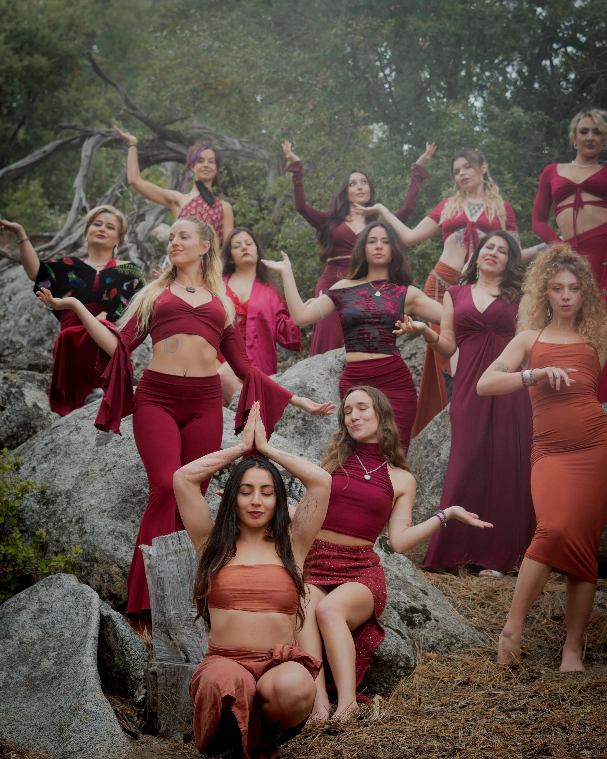 Group of women practicing meditation or yoga outdoors on rocks surrounded by trees, some with eyes closed and hands in prayer or meditative poses.