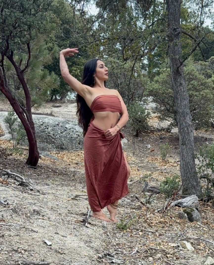 A woman with long dark hair standing barefoot in a forest, wearing a rust-colored wrap top and matching skirt, posing with one arm raised above her head and the other hand resting on her waist.