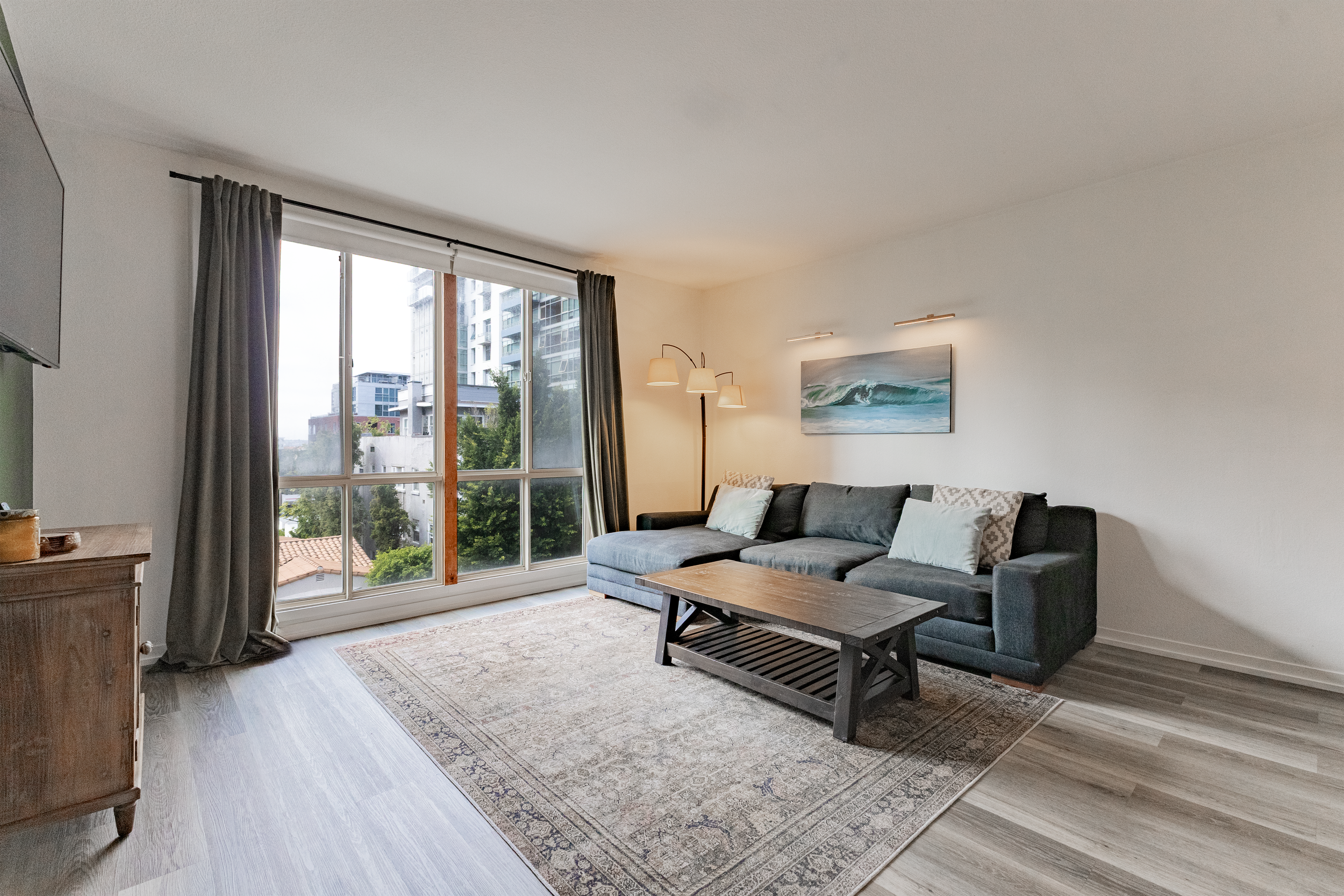 Living room with a large window, gray sectional sofa with pillows, wooden coffee table, painting of waves on the wall, lamps, and a wooden sideboard near the window.
