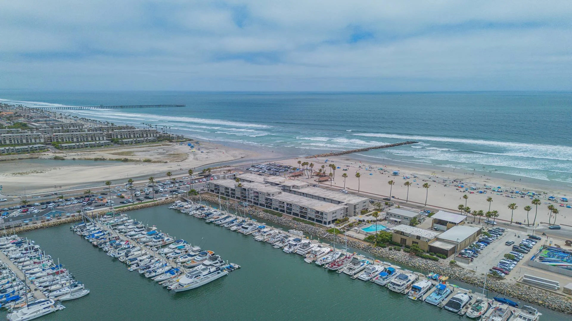 Aerial view of a coastal marina with boats docked in the foreground, a parking lot, a beach with umbrellas, and the ocean with waves in the background, under a partly cloudy sky.