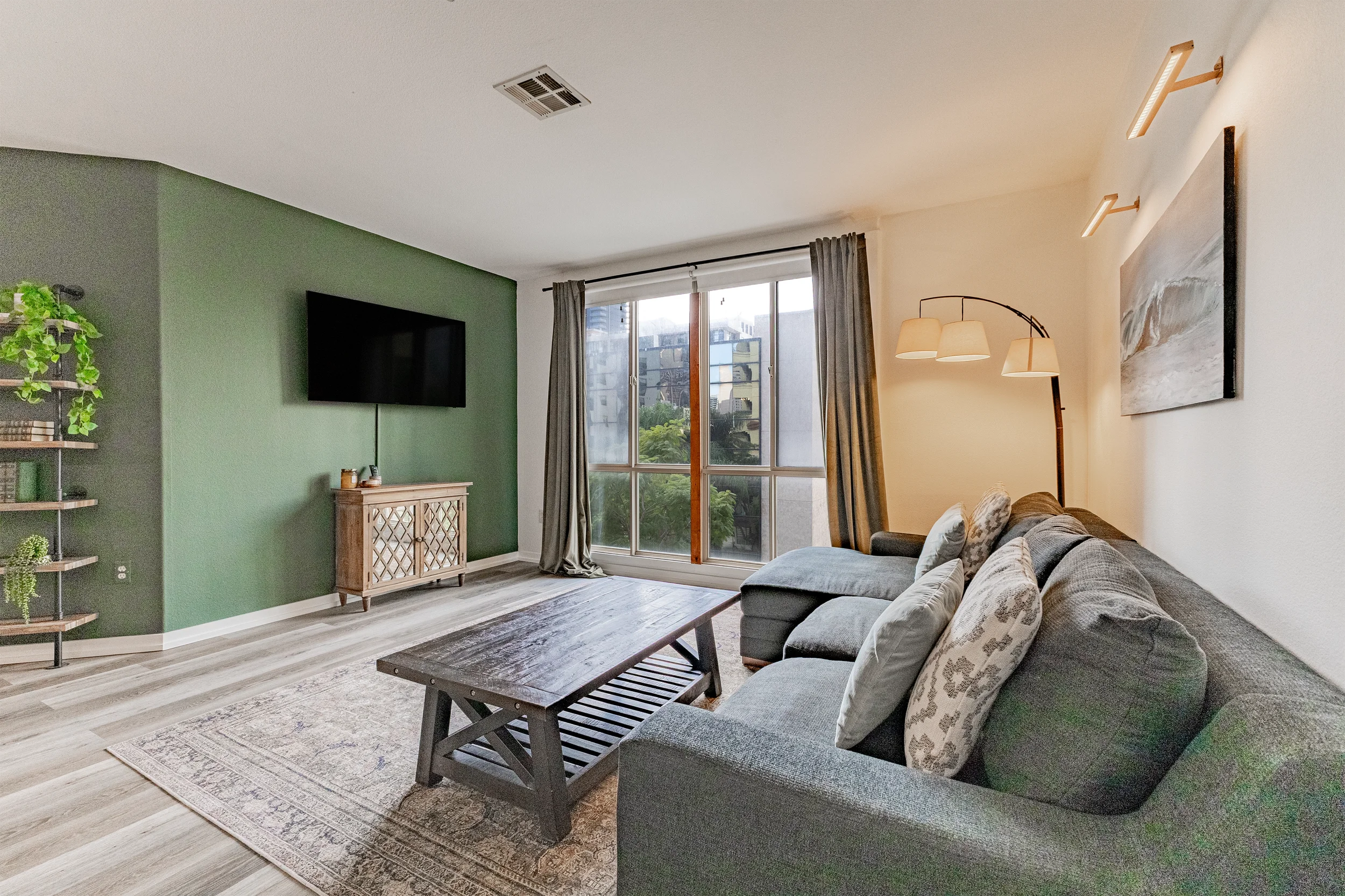 Living room with gray couch, decorative pillows, and a wooden coffee table, next to a large window with curtains, a floor lamp, a painted wall, and a television on a green accent wall.