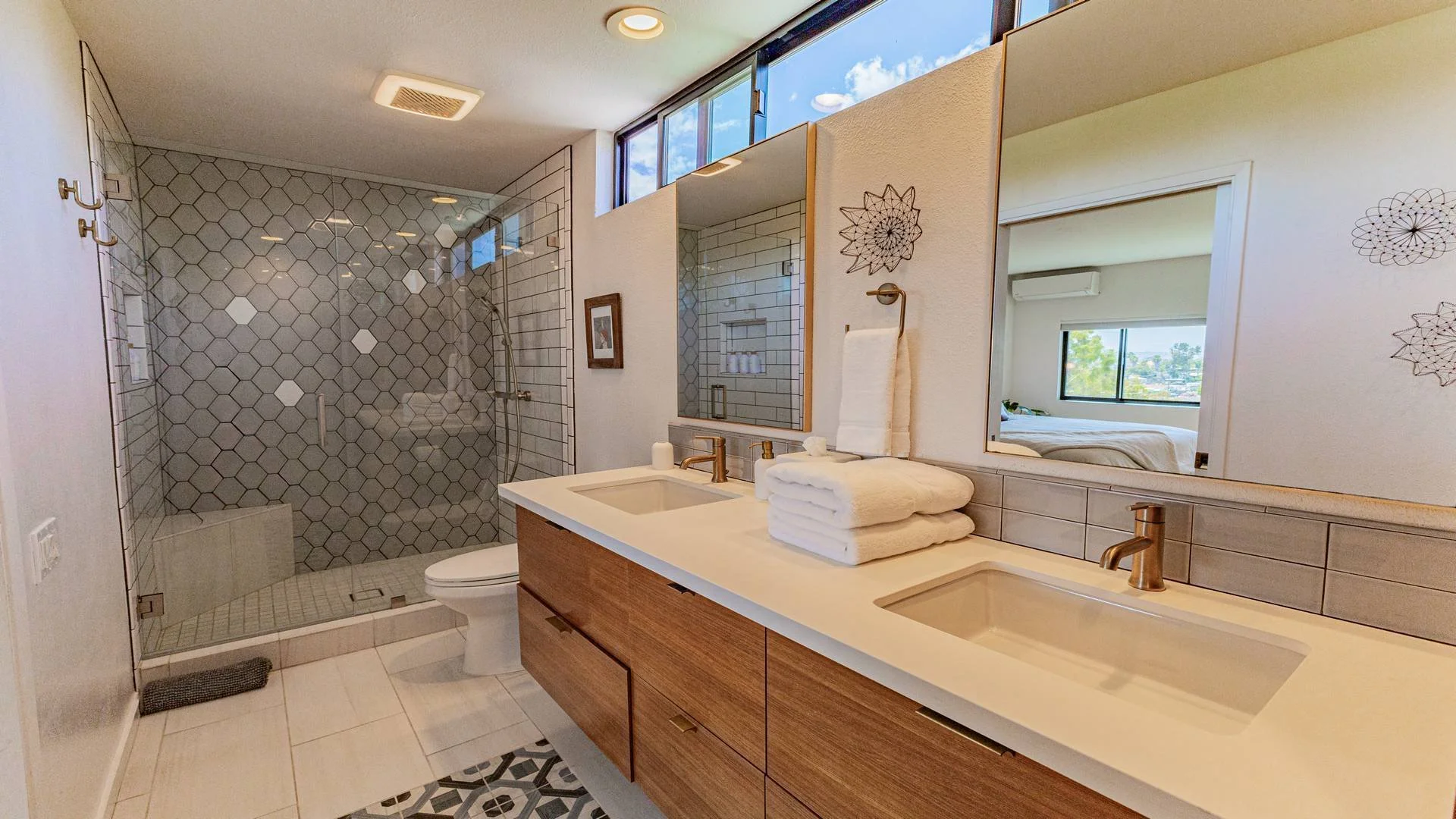 Modern bathroom with double vanity, large mirror, tiled shower, and a view into a bedroom with a large window and bed.