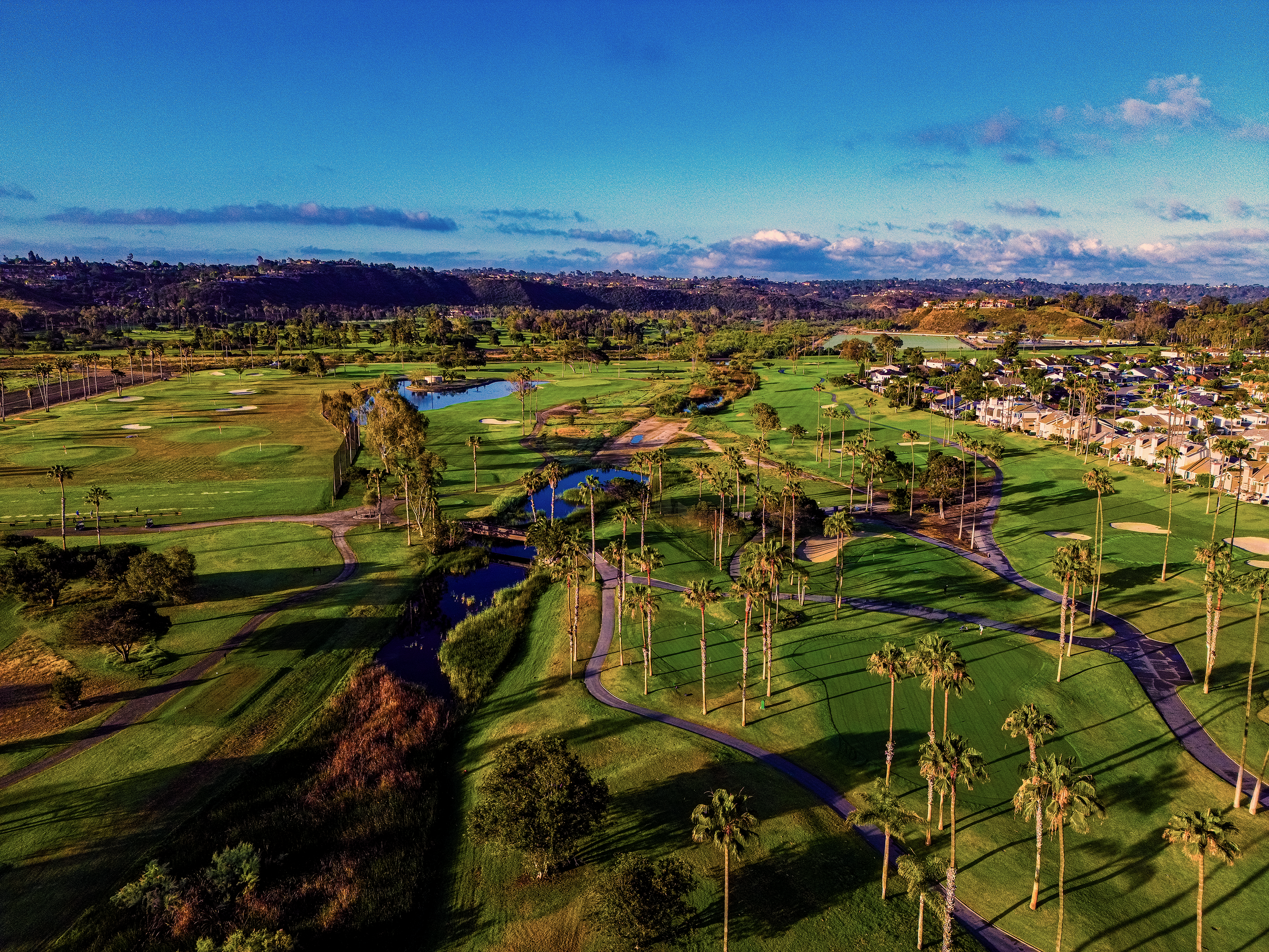 Aerial view of a golf course with green fairways, water hazards, and tall palm trees, with residential houses in the background under a blue sky with some clouds.