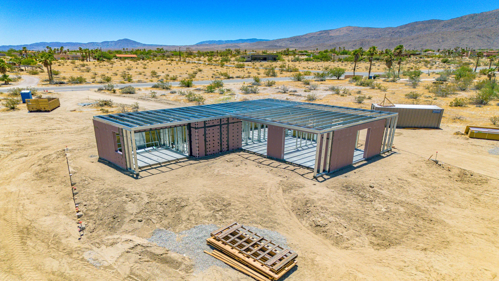 Under-construction house in a desert landscape with mountains in the background. The structure has a metal frame and pink insulation panels, with some walls partially built. Construction materials and containers are on the sandy ground surrounding the site.