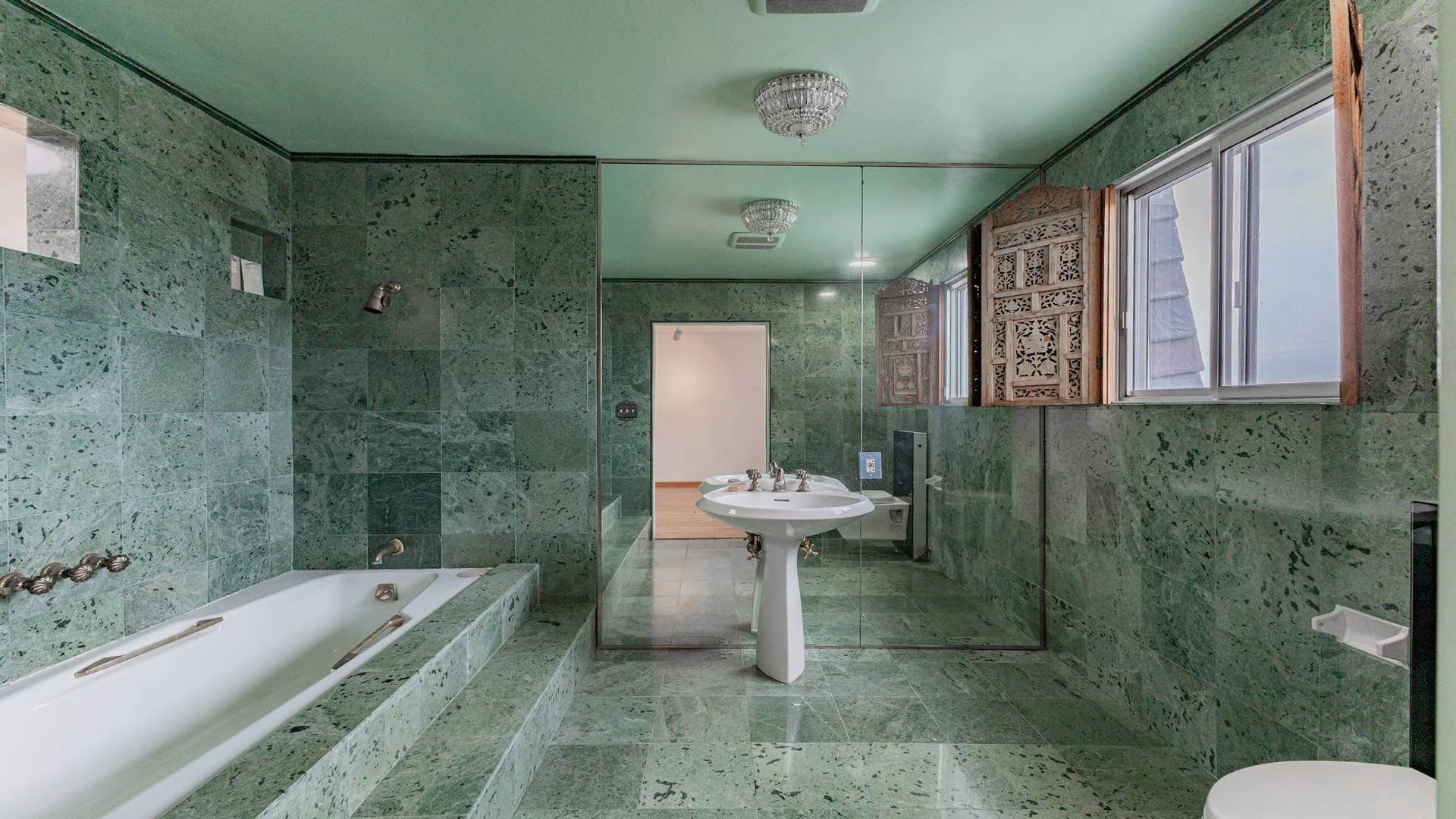 Bathroom with green marble tiles, bathtub, white sink, mirror, large window with wooden lattice, ceiling lights, and a textured ceiling.