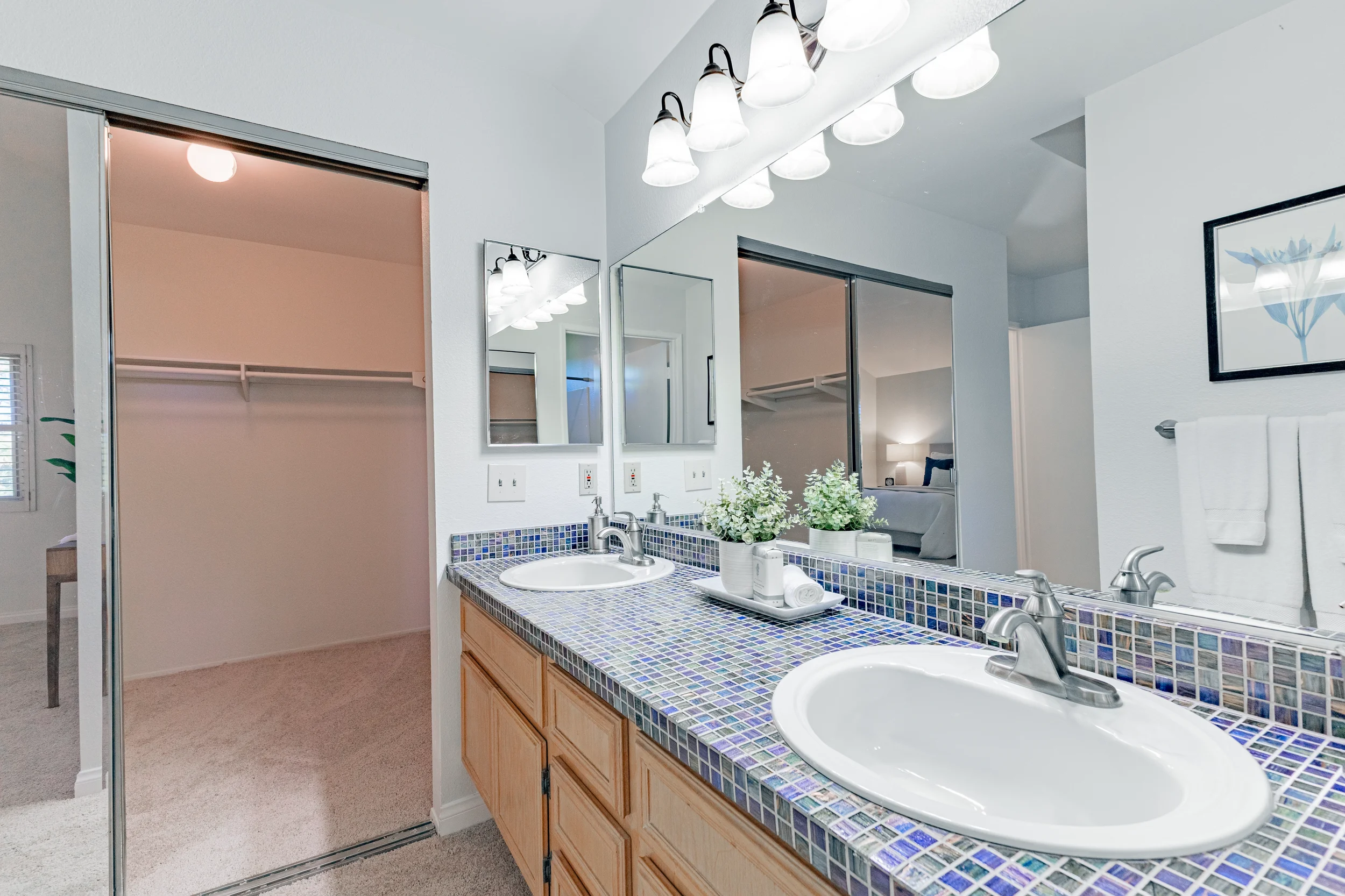 Double bathroom vanity with mosaic tile countertop, two sinks, and mirrors, adjacent to a walk-in closet with carpeted floor, in a bedroom apartment.