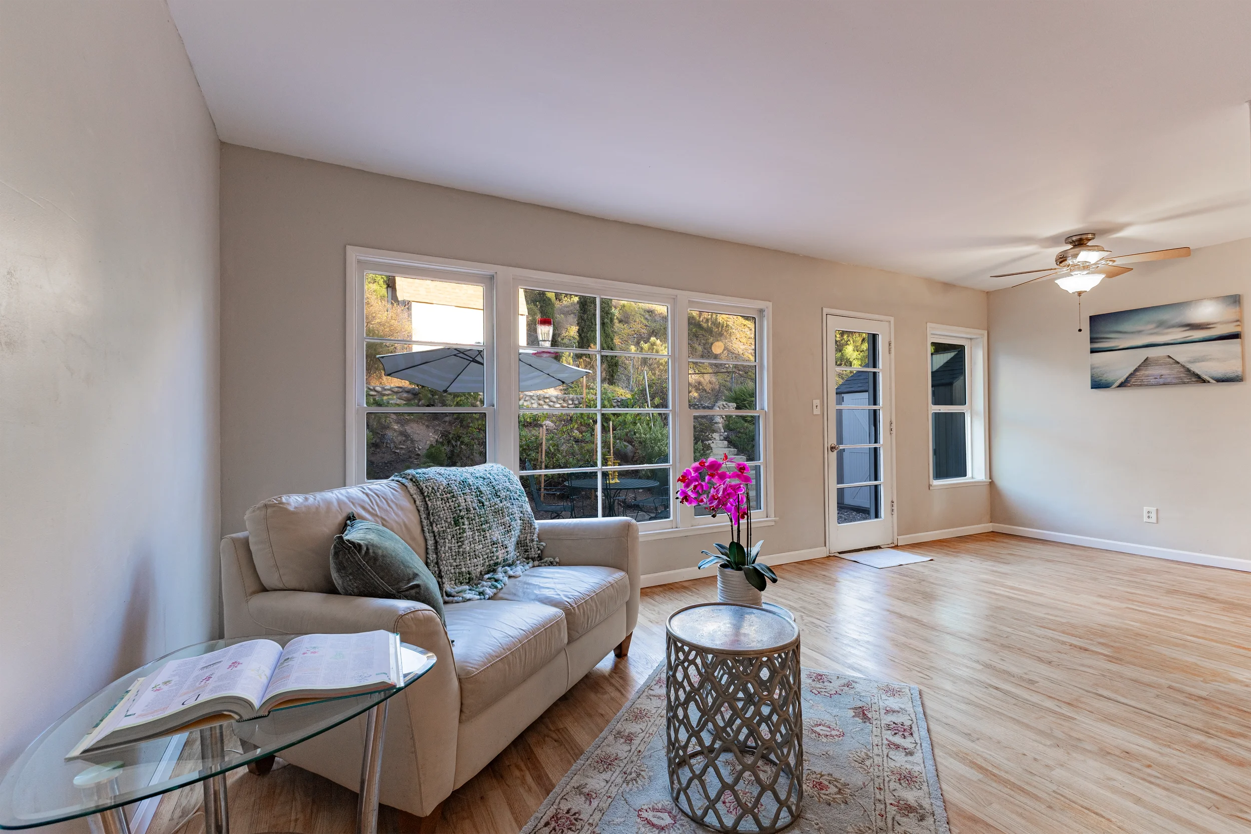 Living room with a white sofa, table with an open book, pink orchid in a pot, light wood flooring, large windows, and a ceiling fan.
