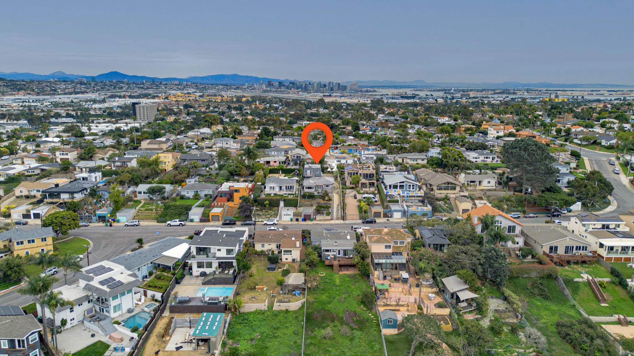 Aerial view of a residential neighborhood with various houses, streets, and greenery, overlooking a city skyline and hills in the distance.