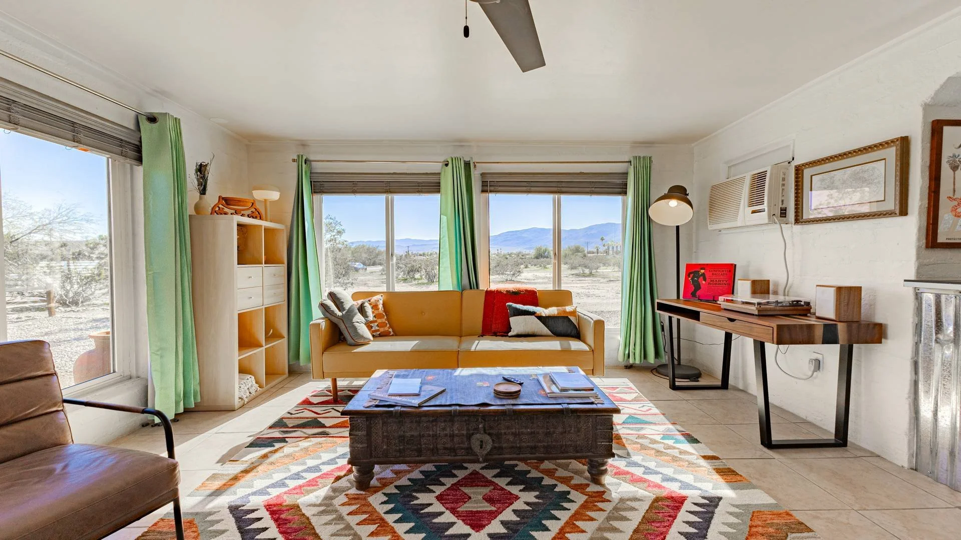 Living room with large windows showing desert landscape and mountains, yellow sofa with pillows, wooden coffee table, multicolored geometric rug, bookshelf, wall art, and radio on a wooden console.