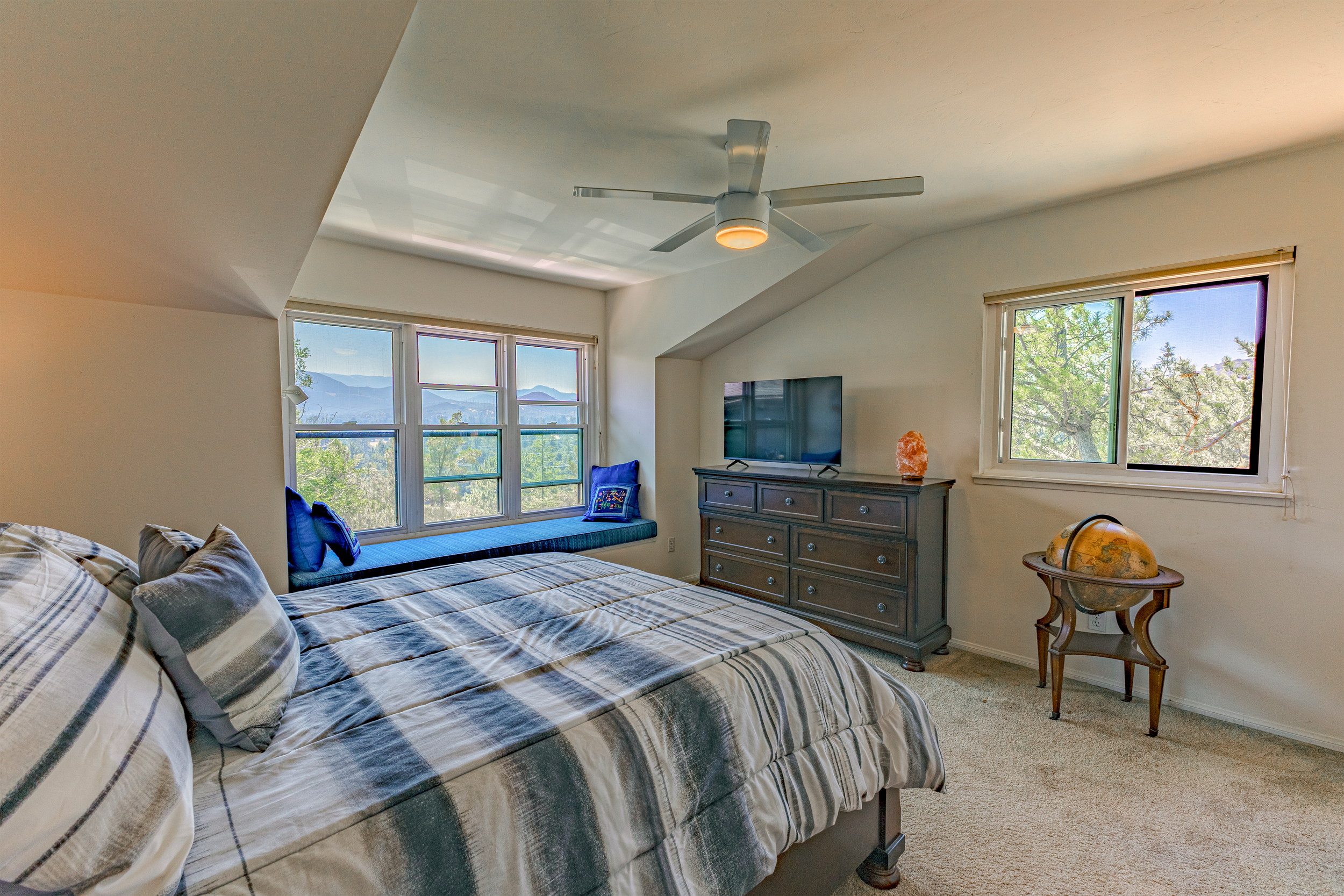 Bedroom with a bed, window seat, dresser, and two windows showing a scenic view of trees and mountains.