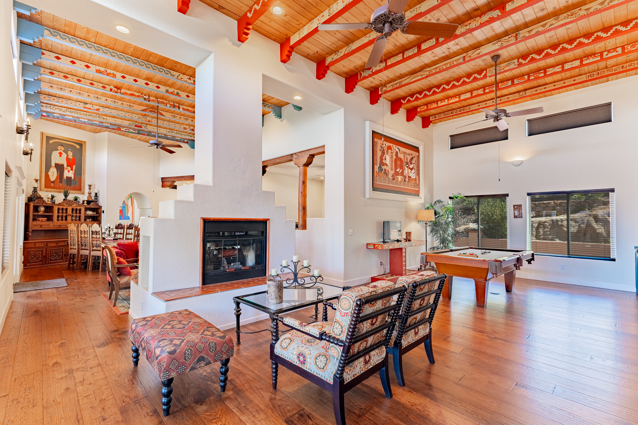 Living room with wood floor, a pool table, a fireplace, and a sitting area. Decor includes framed artwork, a lamp, and assorted furniture and plants. The ceiling shows exposed wooden beams with decorative patterns.