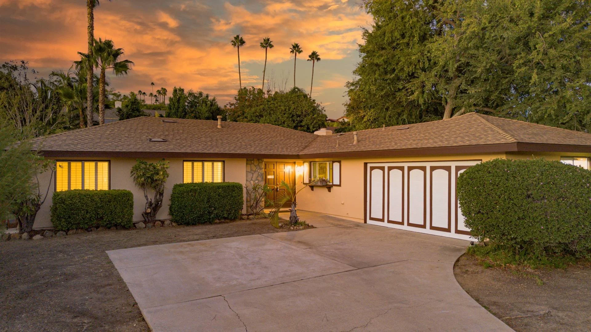 Single-story house with a brown shingle roof, front yard with trimmed bushes, illuminated windows, and a garage door with dark brown framing. The house is surrounded by tall palm trees and lush green trees, with a colorful sunset sky in the backgroun