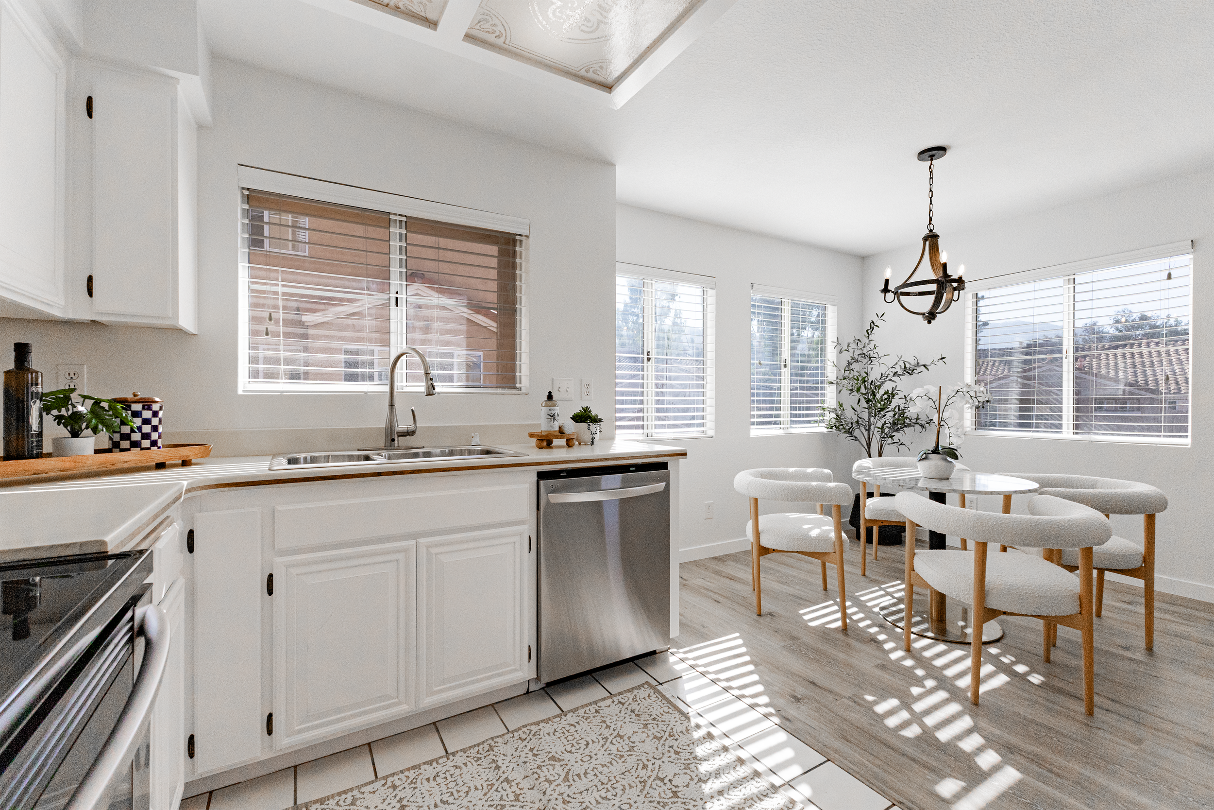 Bright, modern kitchen and dining area with white cabinets, stainless steel dishwasher, and a round dining table with four cushioned chairs, illuminated by natural light from large windows.
