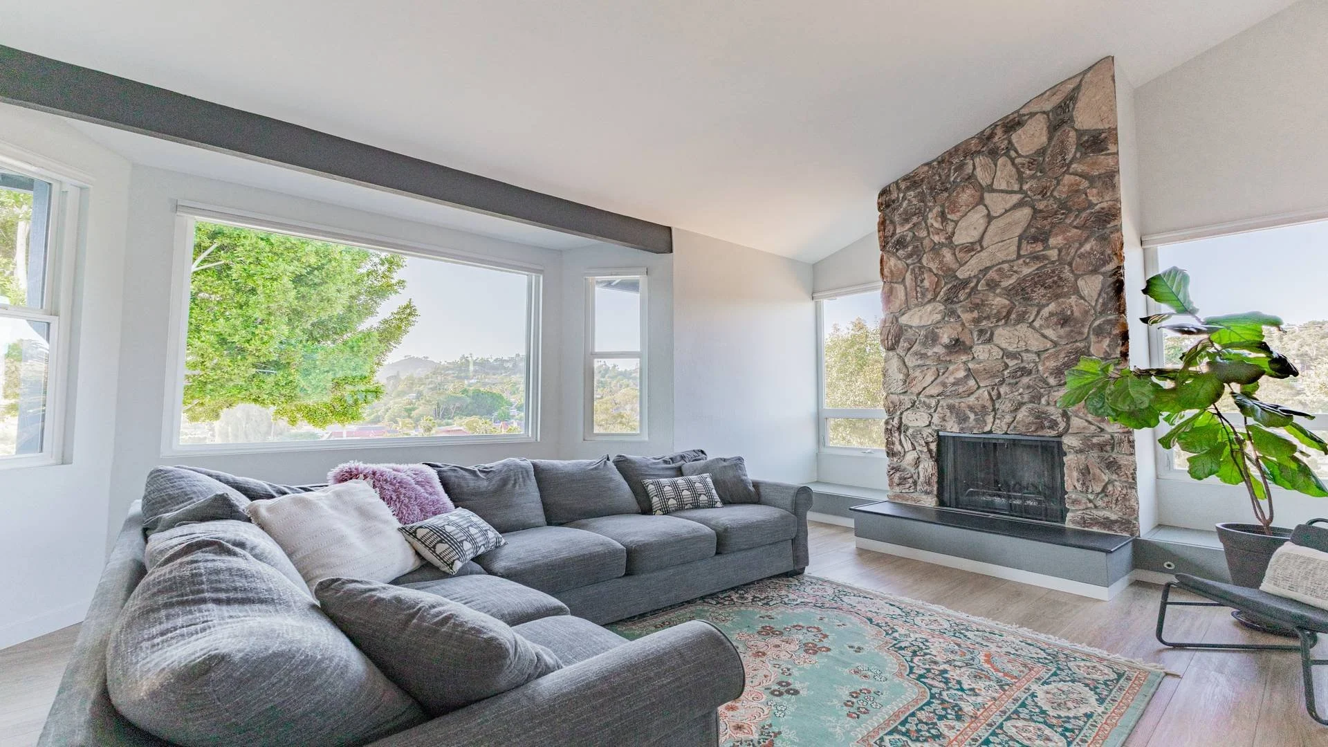Living room with large windows showing trees and landscape, gray sectional sofa with pillows, a colorful area rug, a stone fireplace, and a potted plant nearby. Luxury real estate photography in La Mesa. 
