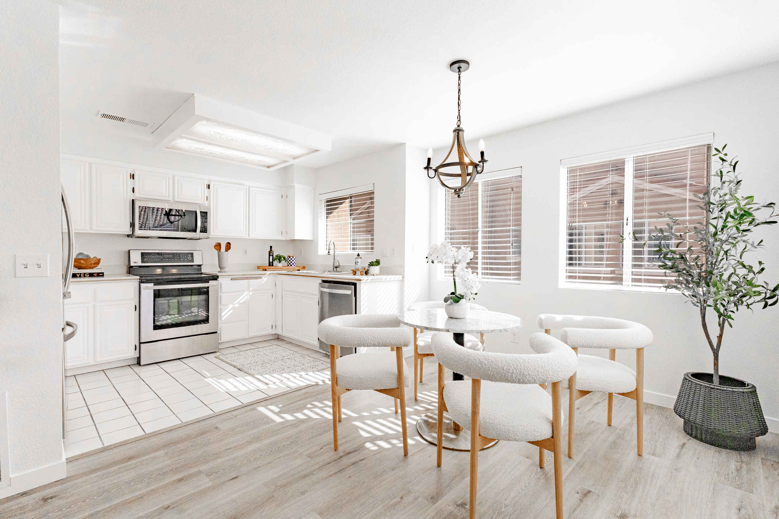 Bright, modern kitchen and dining area with white cabinets, stainless steel appliances, a round marble table with four white upholstered chairs, a large potted plant, and large windows with blinds letting in natural light.