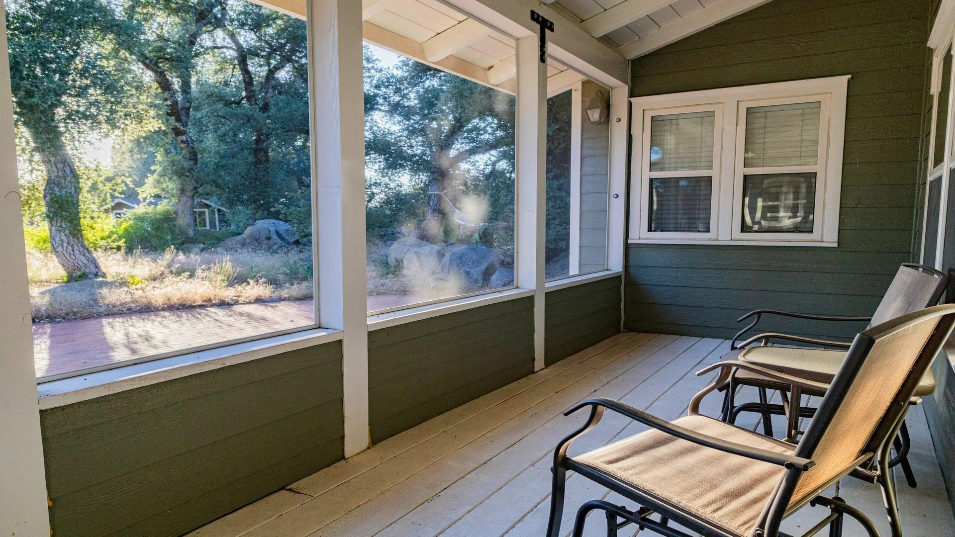 A screened porch with beige wooden floorboards, green siding, and white trim. Four metal and fabric chairs are arranged on the right side, facing the view of trees and rocks outside. The porch has a two-pane window and a beige light fixture on the wa