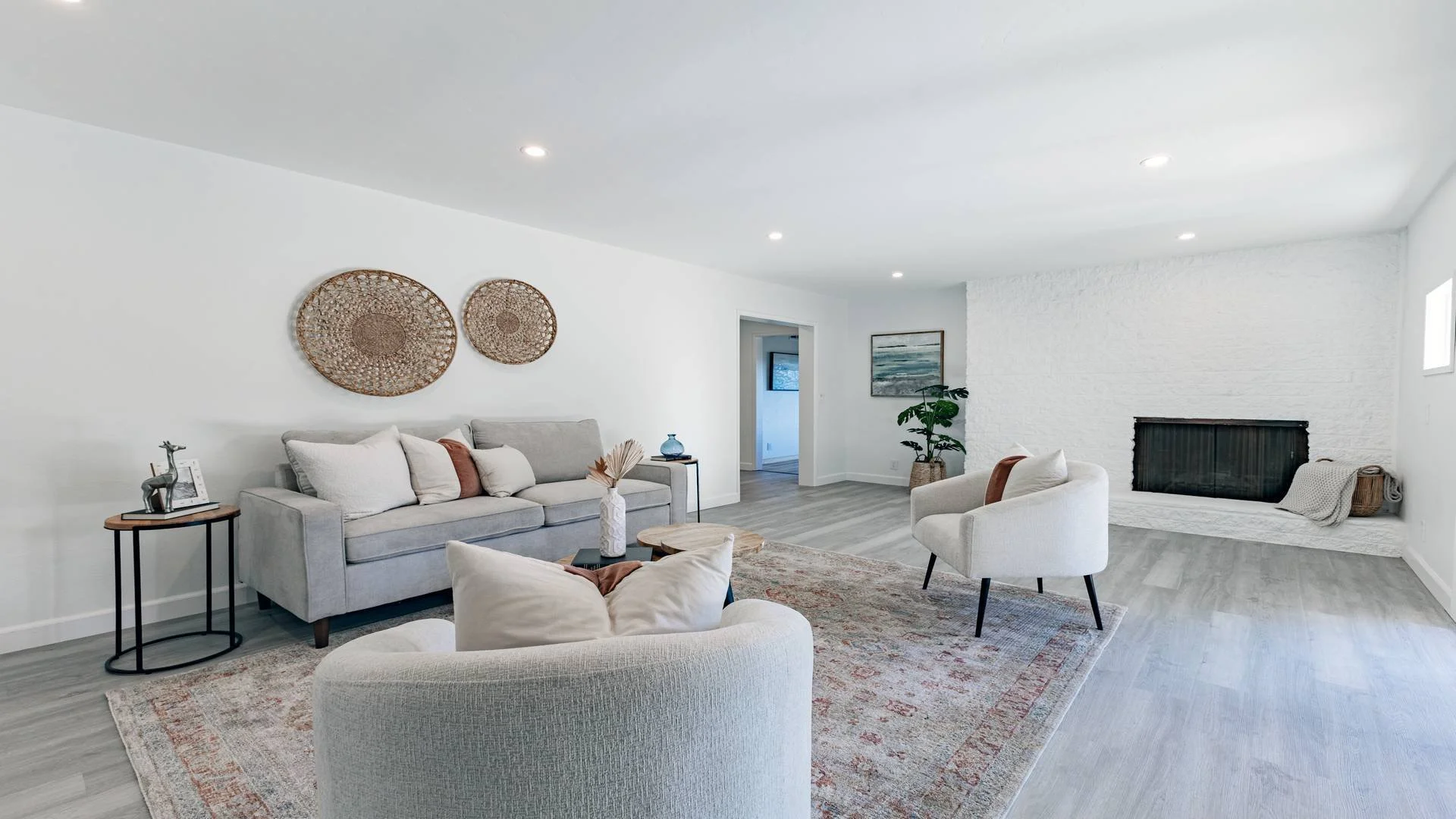 Bright living room with white walls and light wood flooring, decorated with woven wall art, a gray sofa, white armchair, potted plant, fireplace, and cozy decor. Real estate photography of a property in El Cajon. 