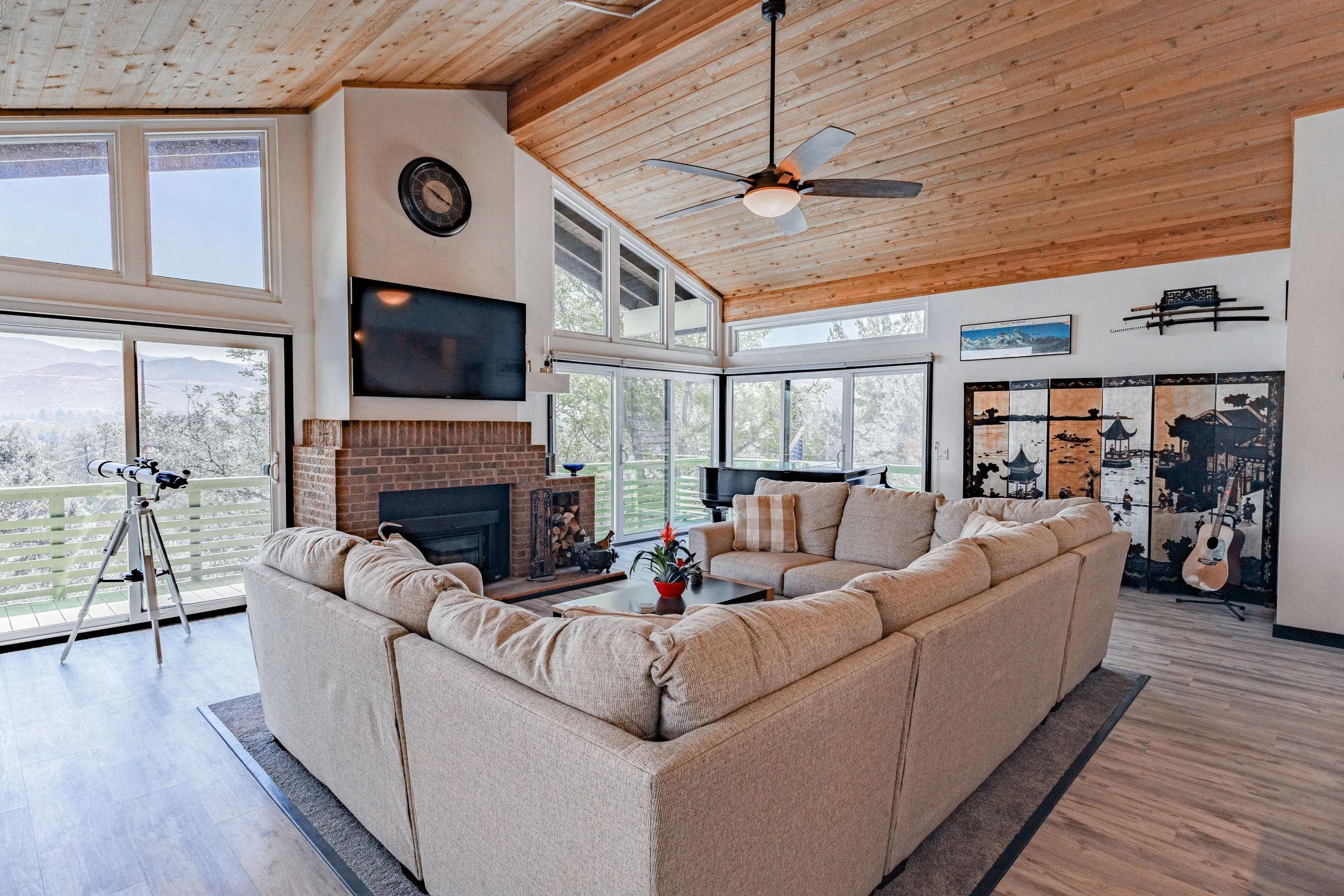 Living room with large sectional sofa, brick fireplace, flat-screen TV, telescope, potted plant, and Asian-style folding screens, with windows showing trees and mountains outside.