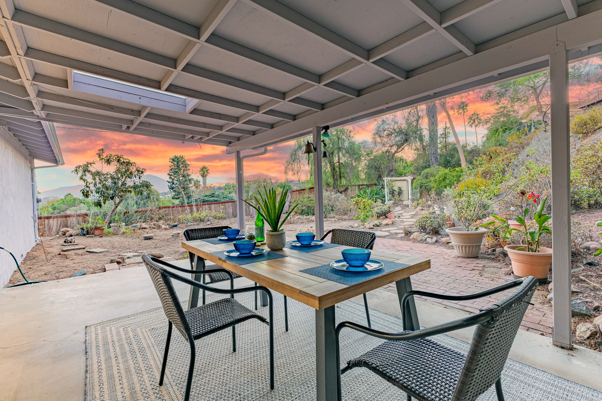 View of a backyard patio with a wooden dining table set for four, blue bowls and a potted plant, overlooking a garden at sunset with trees and colorful sky.