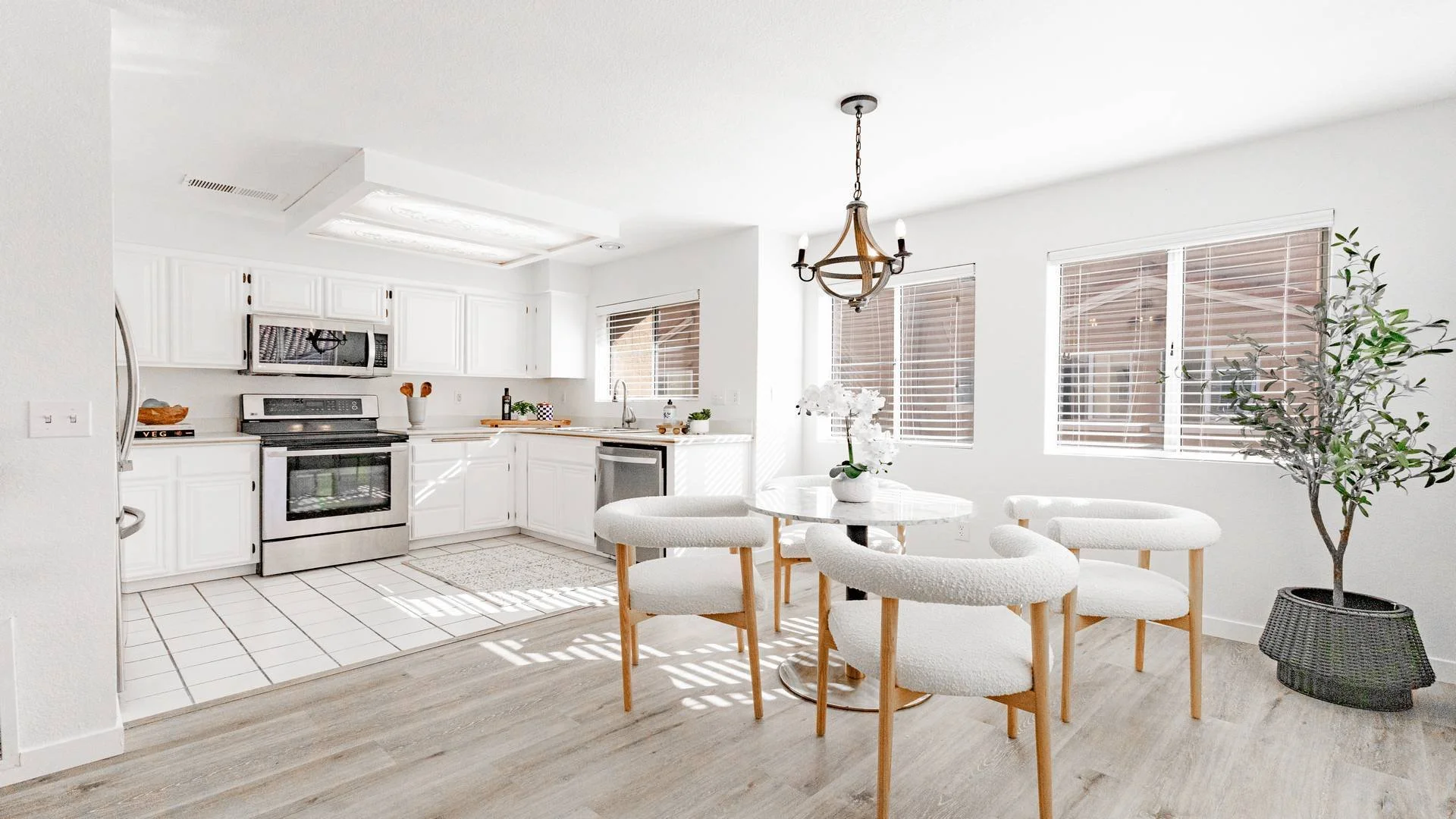 Bright kitchen and dining area with white cabinets, stainless steel appliances, a round white table, white chairs, potted plants, and multiple windows with white blinds in an El Cajon home. 