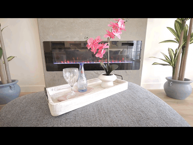 A rectangular tray with a glass, a blue vase with pink flowers, and a white vase with pink flowers, placed on a gray cushion in front of a modern fireplace with a concrete surround, flanked by two large green potted plants.