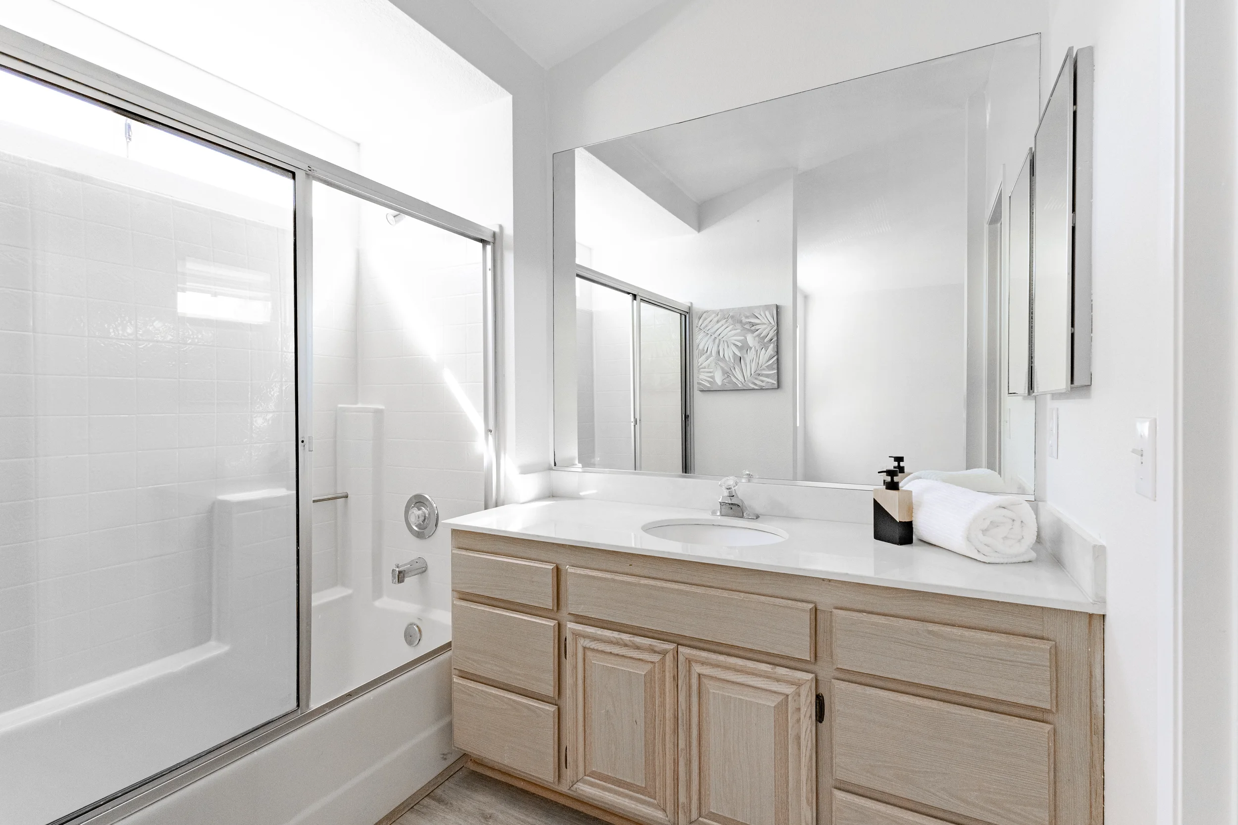 Bathroom with a white shower with glass doors, a white vanity with a round sink, a large mirror, a rolled towel, and a black soap dispenser.