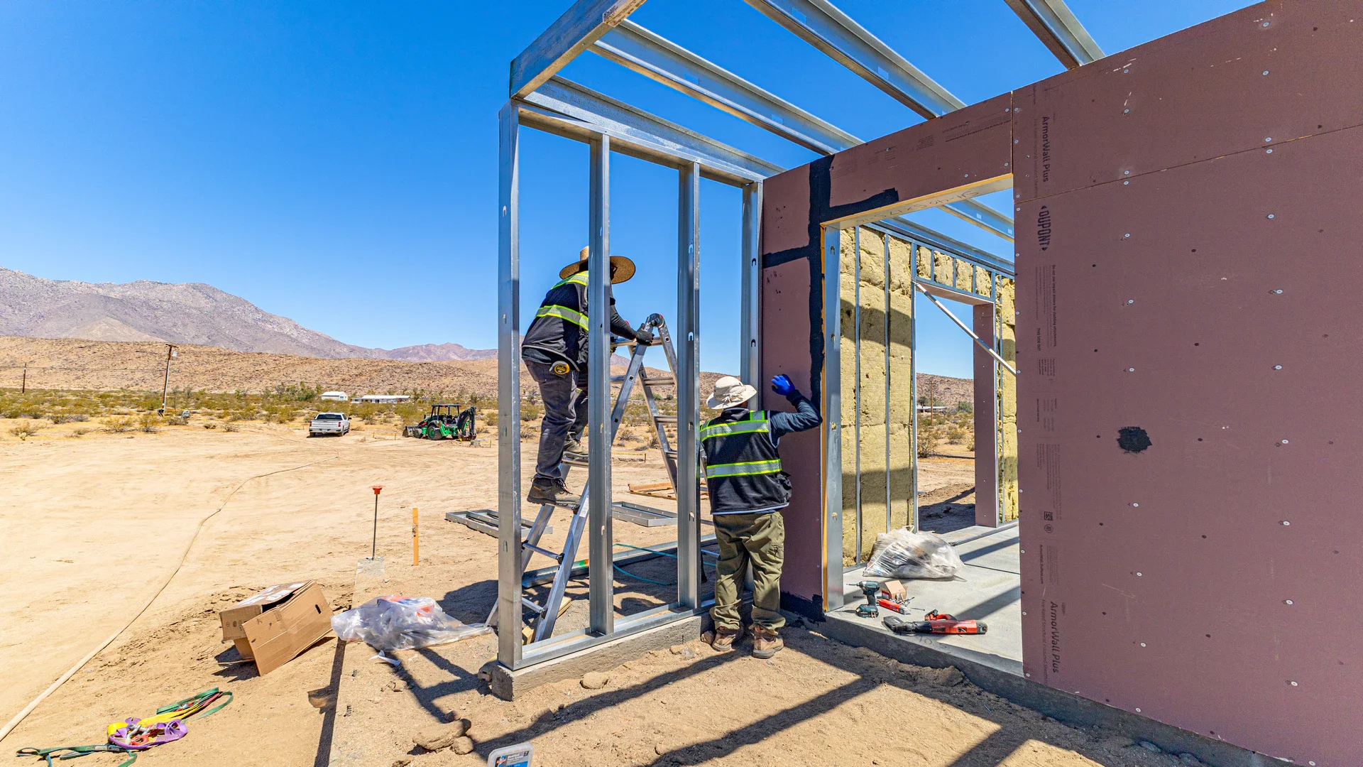 Two workers wearing safety vests and hats assemble a building frame in a desert construction site, with mountains and vehicles in the background.