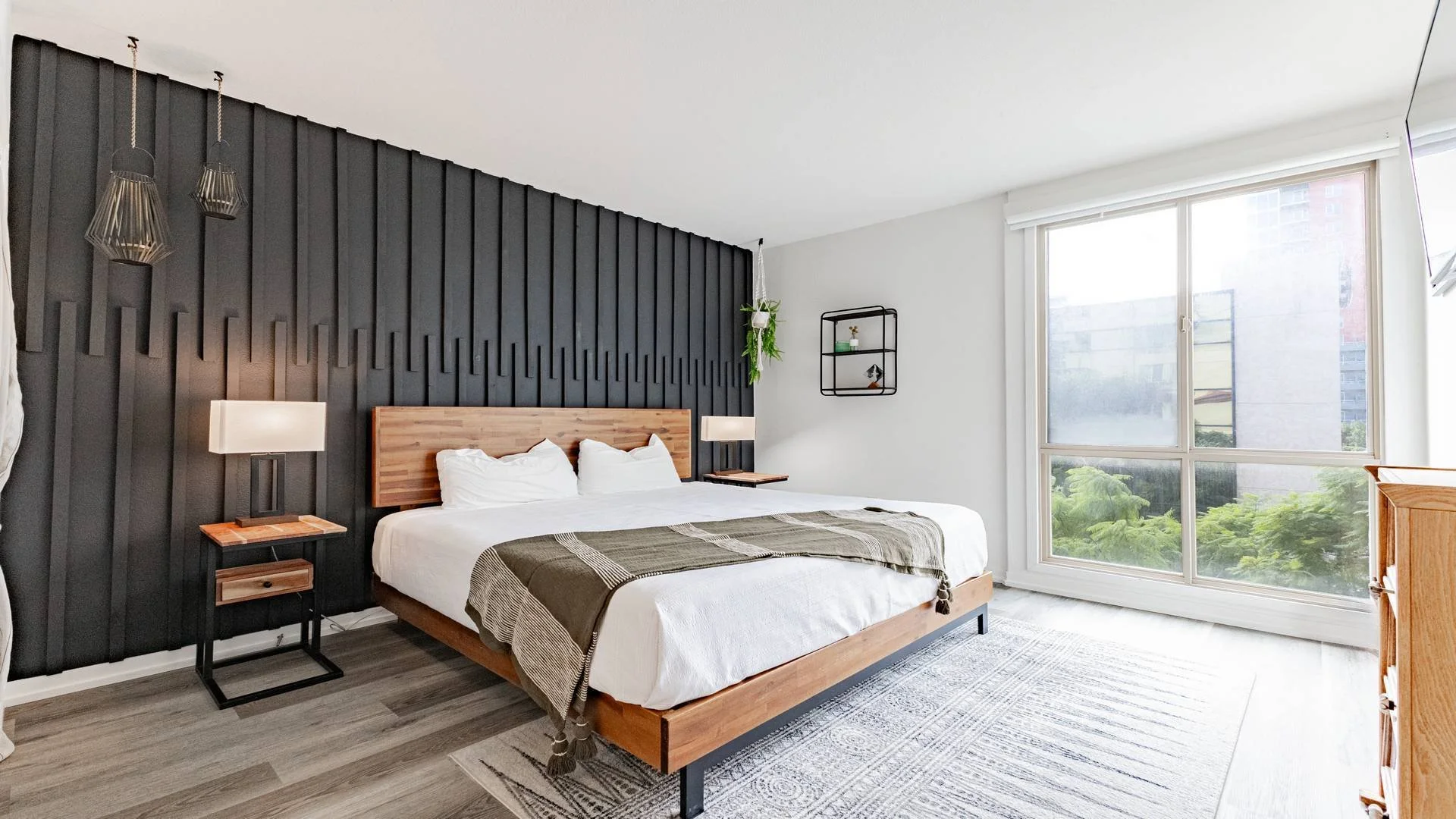 Modern bedroom with black accent wall, wooden bed frame, white bedding, side tables with lamps, large window showing trees and buildings outside, and hanging plants.