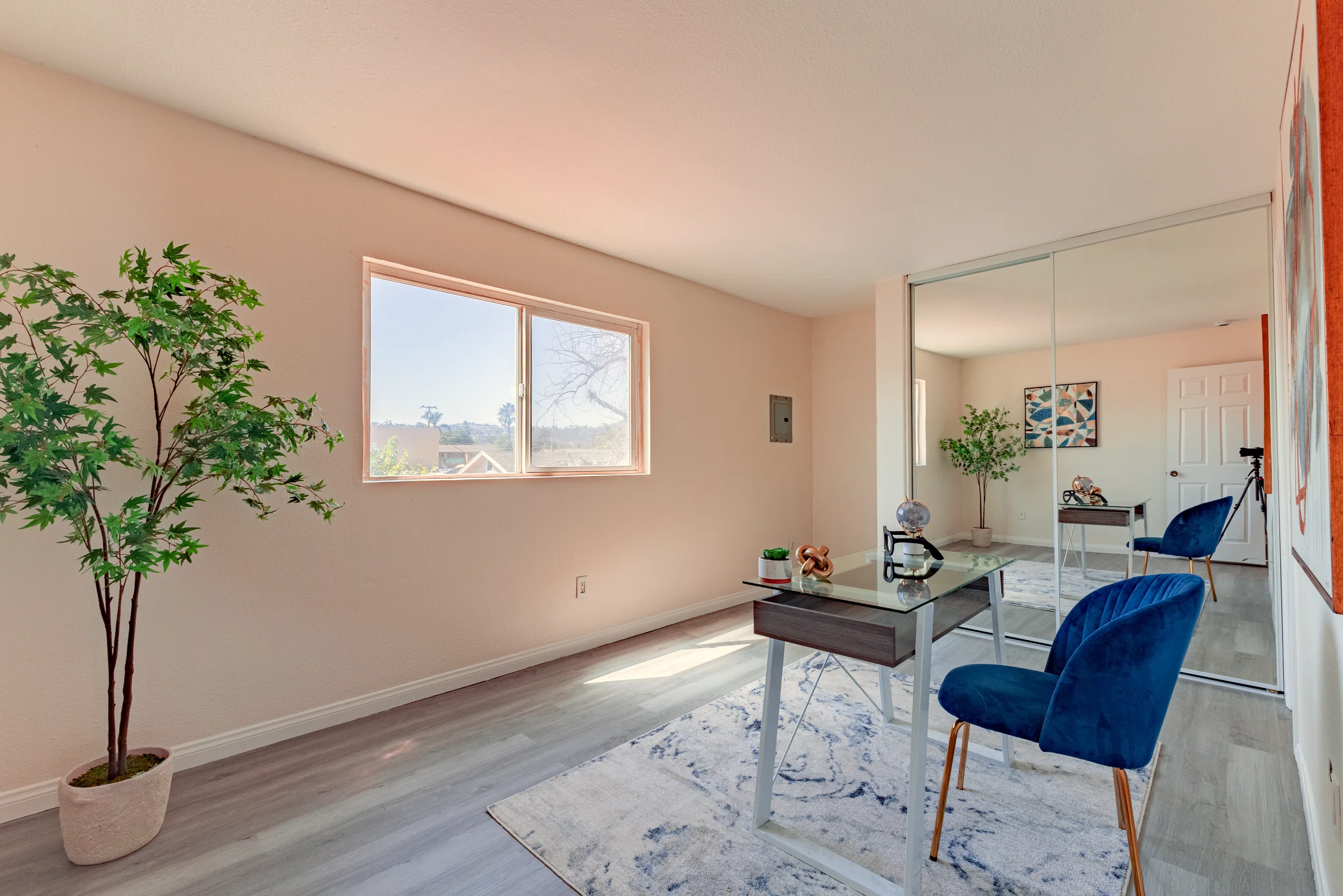 Room with a large window, potted plant, glass desk with blue chair, mirror sliding closet doors, decorative items, and artwork on the wall.