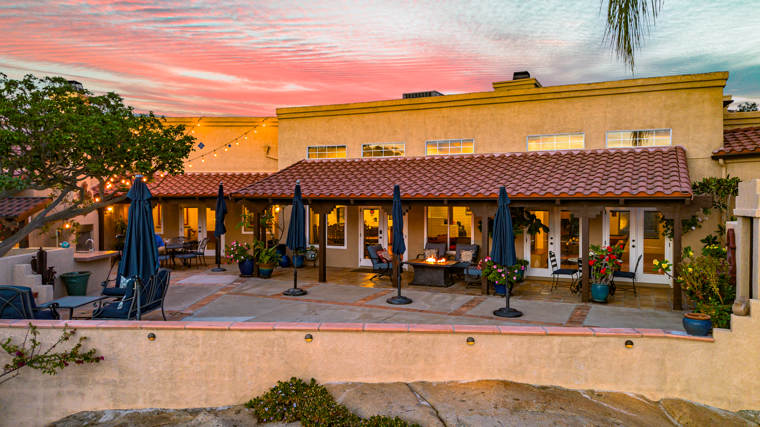 Backyard patio of a house during sunset with outdoor seating, umbrellas, string lights, decorated with potted plants, and a fire pit.