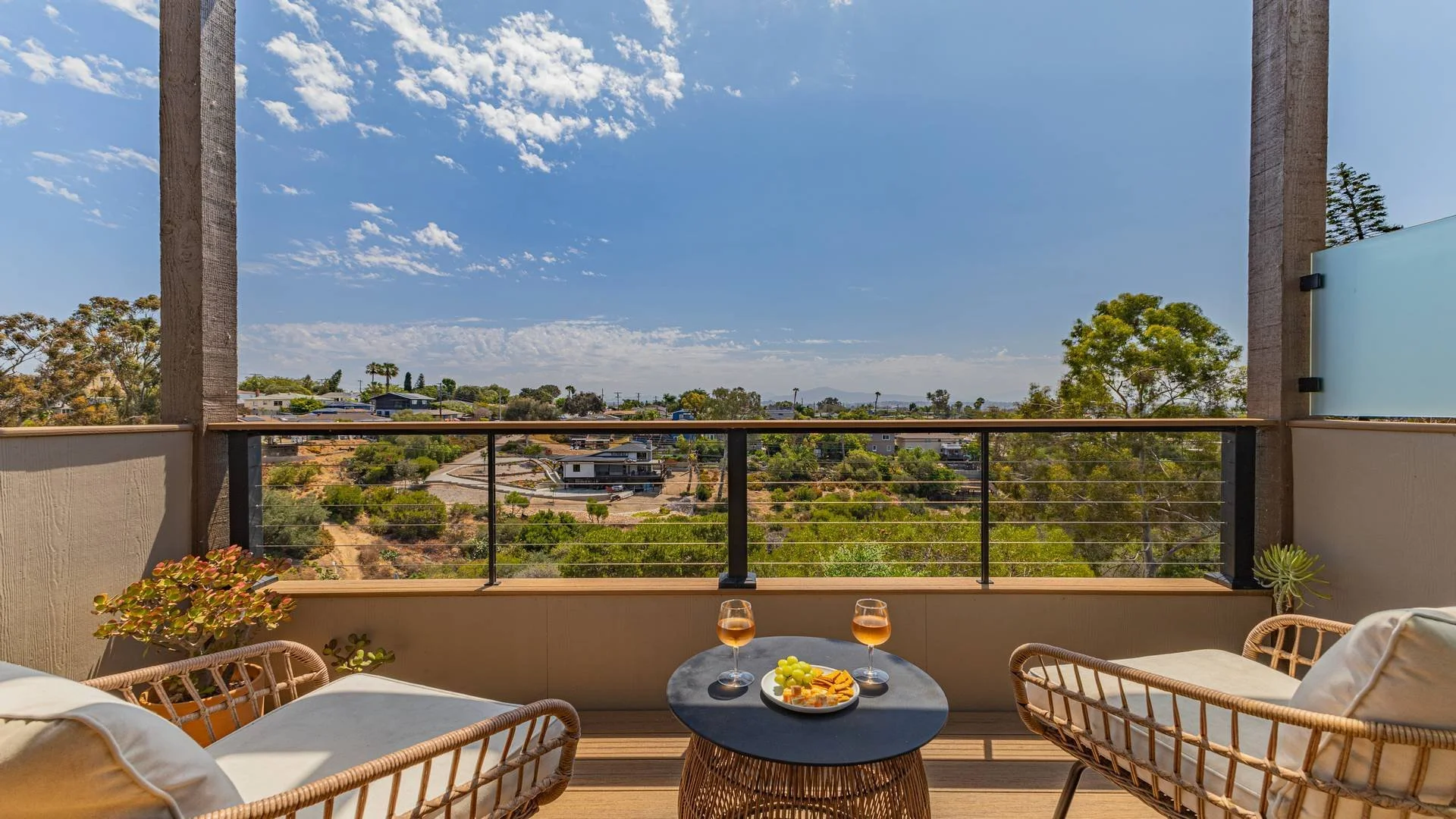Balcony view overlooking suburban neighborhood with trees and houses, blue sky with some clouds, and outdoor furniture with drinks and snacks.