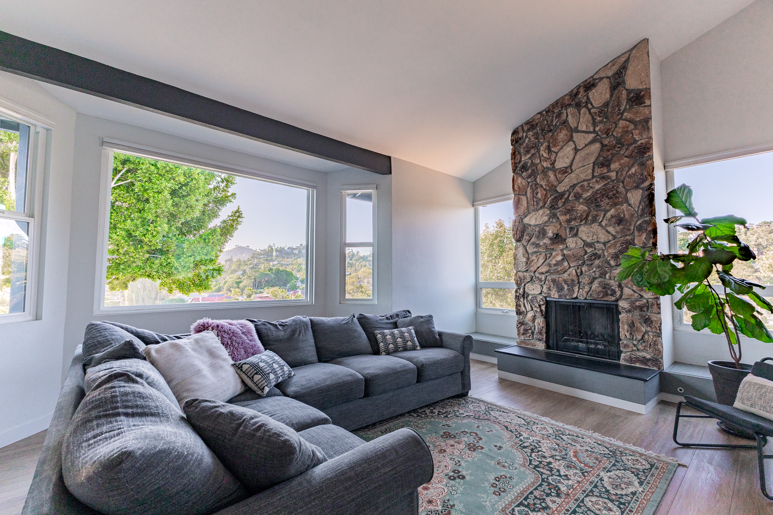 Living room with large windows, a gray sectional sofa with throw pillows, a stone fireplace, a plant, and a patterned area rug.