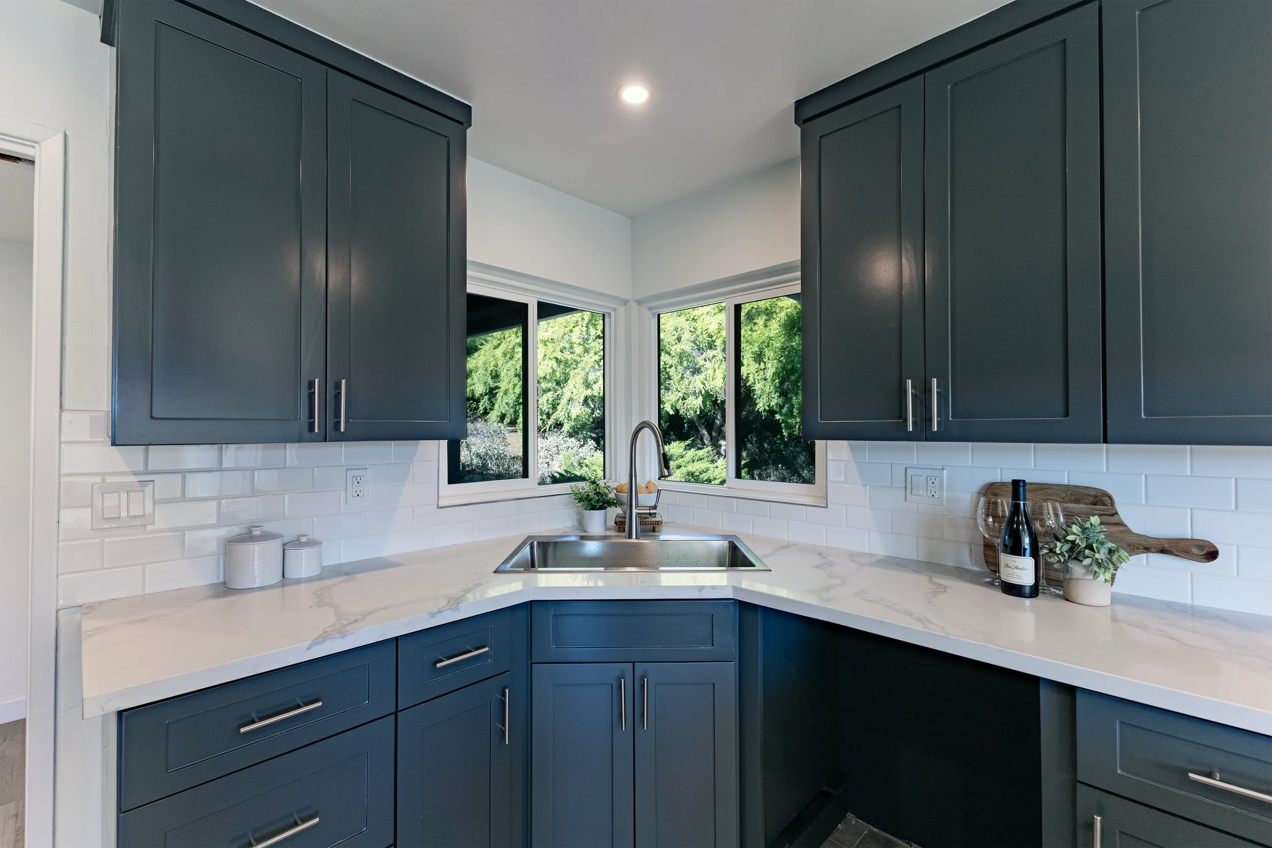 Kitchen with blue cabinets, white marble countertop, white subway tile backsplash, stainless steel sink, and windows showing greenery outside.