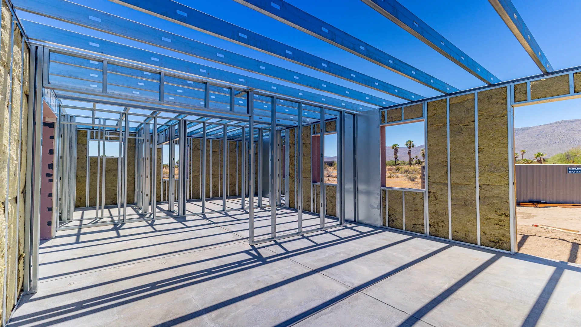 Construction site with metal framing for walls and roof of a building in a desert environment with mountains and palm trees in the background.