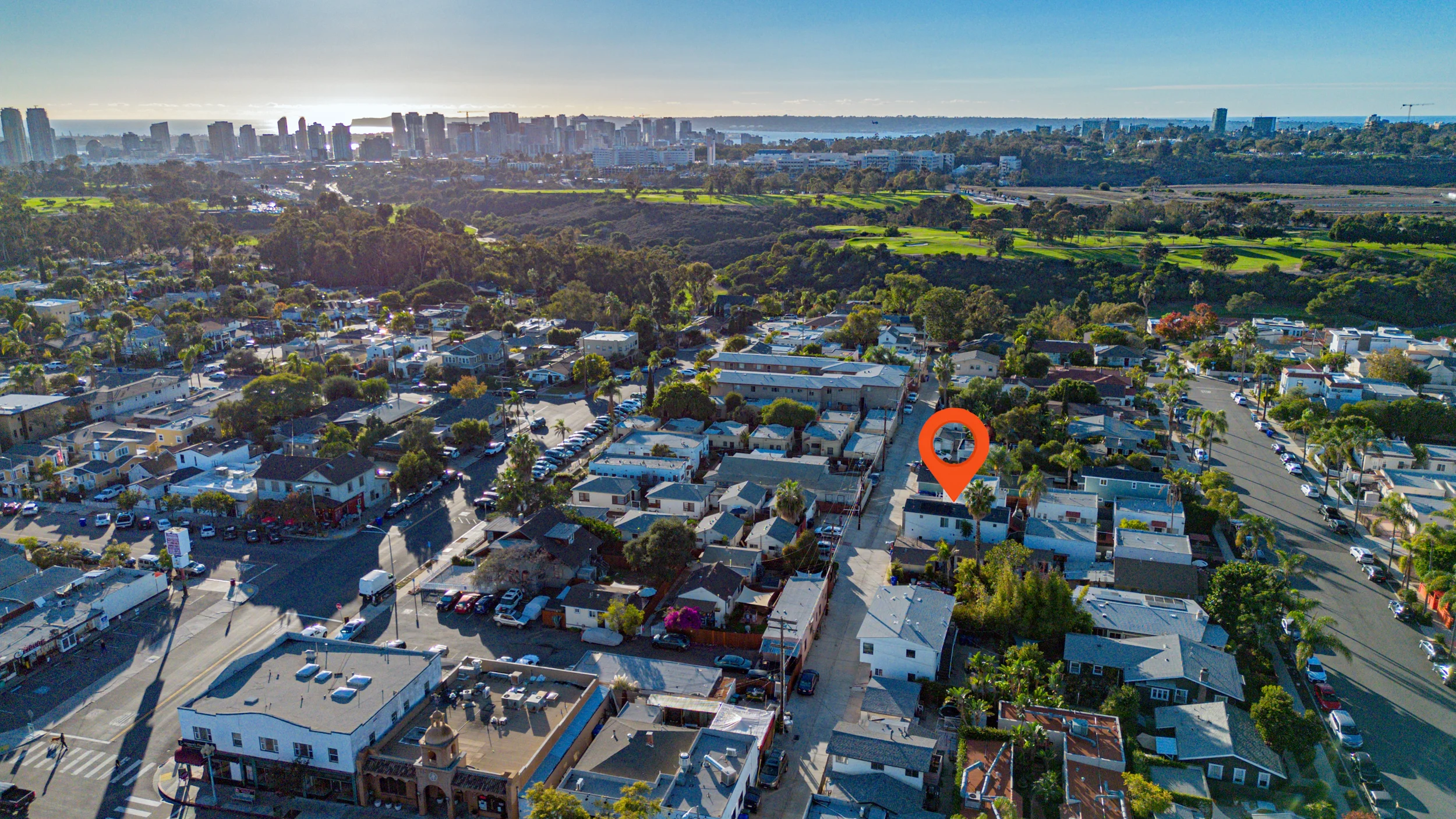 Aerial view of a neighborhood with houses, trees, and streets, with a city skyline and green fields in the background.