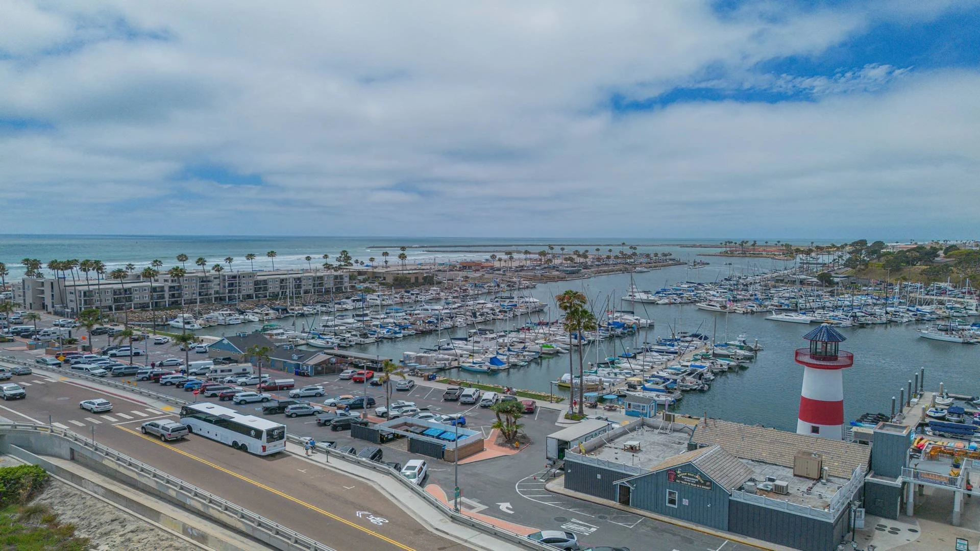 A coastal marina with numerous boats docked, a lighthouse on the right, palm trees, and a cloudy sky.