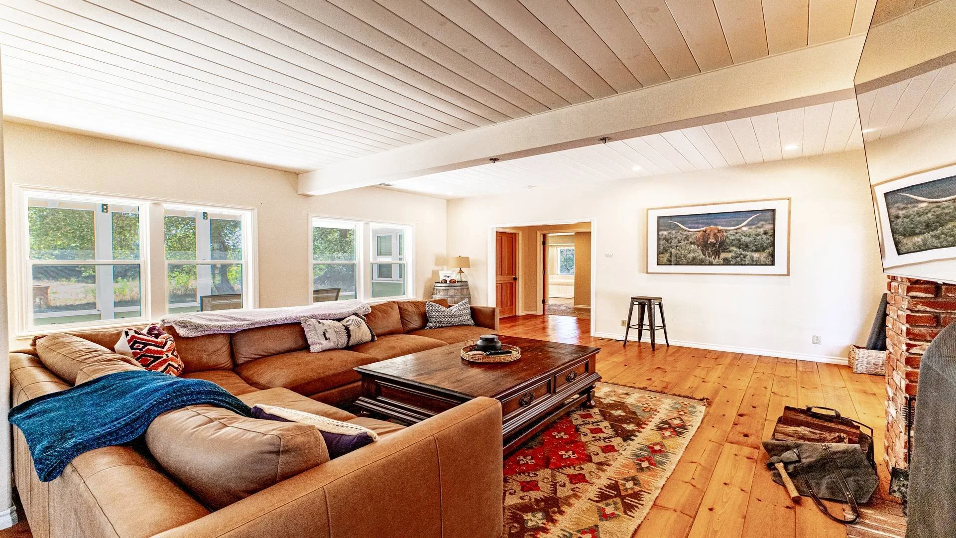 Living room with large brown sectional sofa, wooden coffee table, framed cowboy scene on wall, brick fireplace, three windows, hardwood floors, and various pillows and blankets. Airbnb photography for a property in San Diego County. 