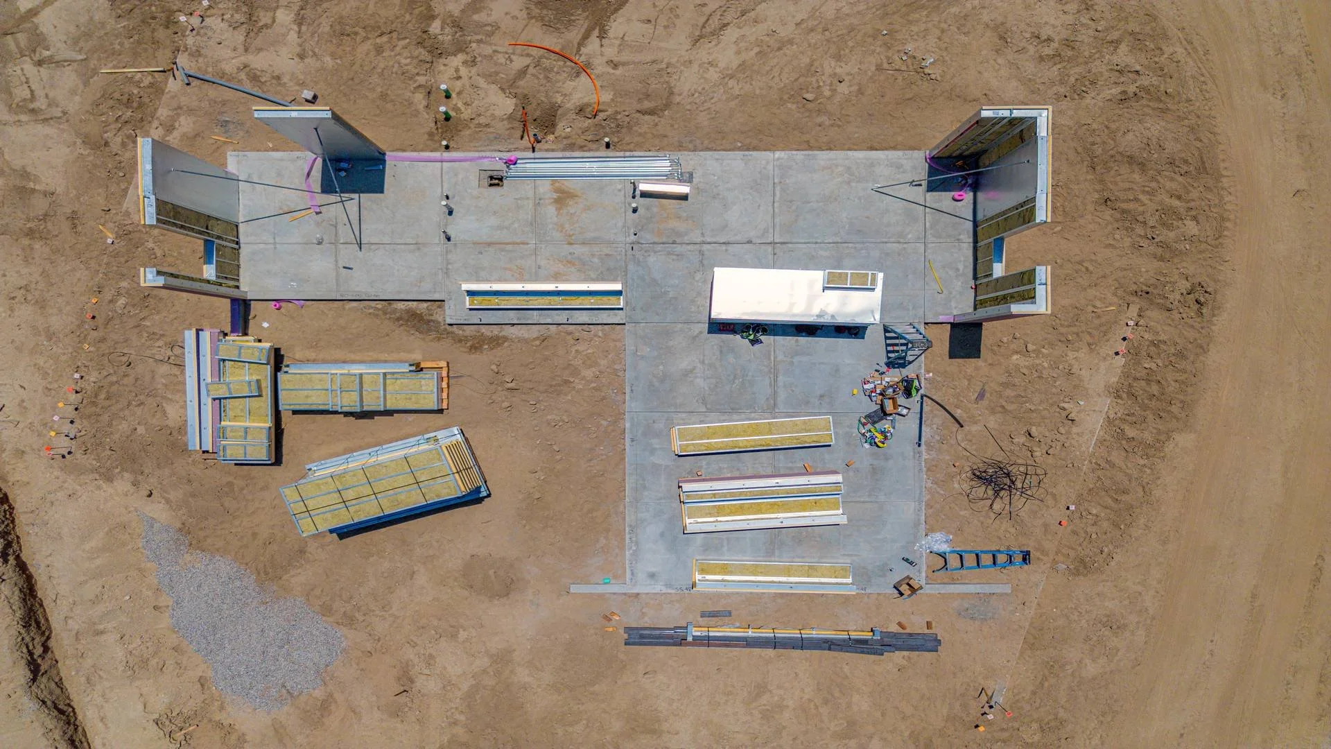 An aerial view of a construction site with a concrete slab foundation surrounded by dirt, scattered construction materials, and equipment, with partially framed walls and metal structures.