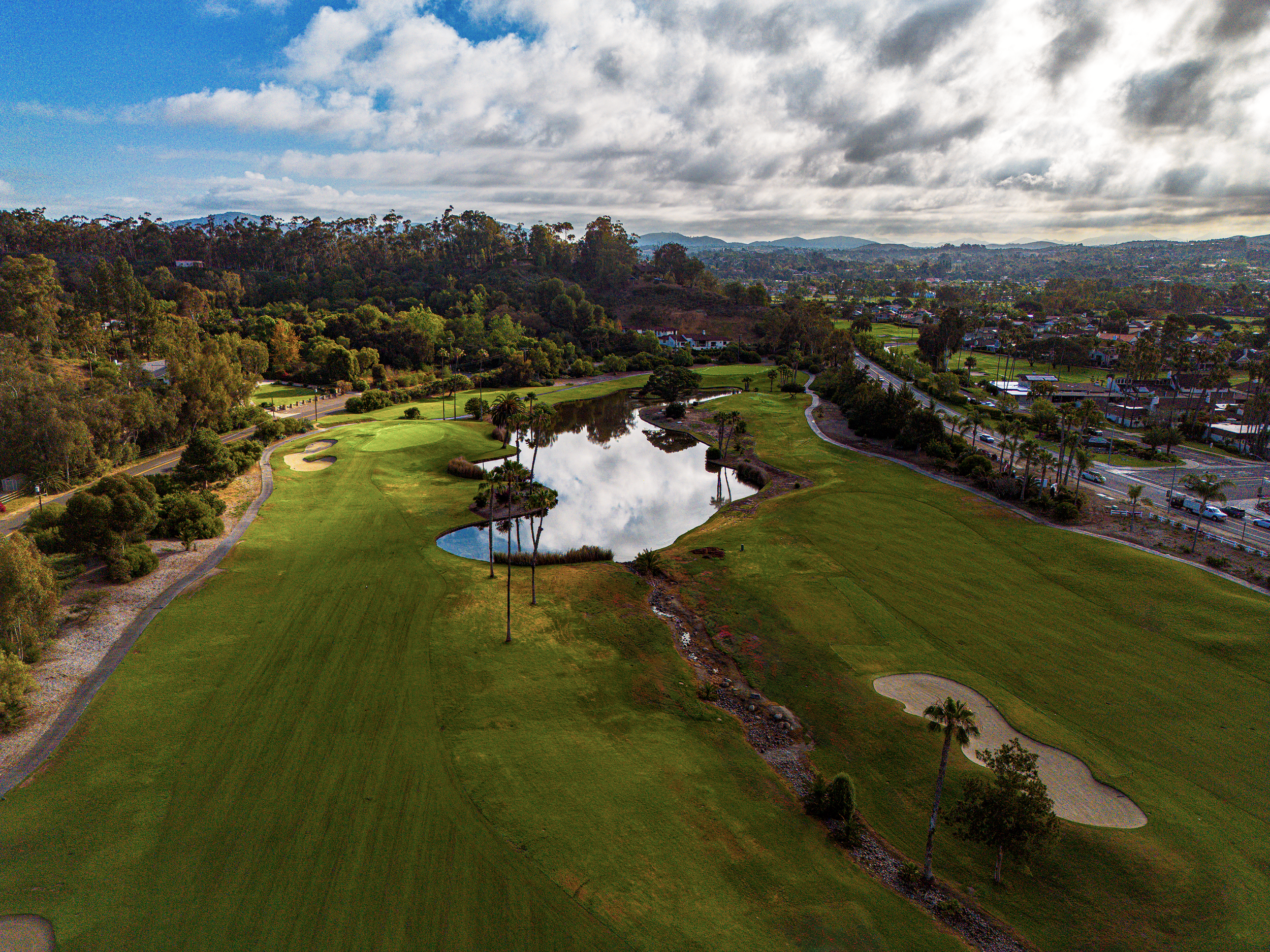 An aerial view of a golf course with green grass, a pond, and surrounding trees. In the background, there are residential houses and a town, with hills under a partly cloudy sky.