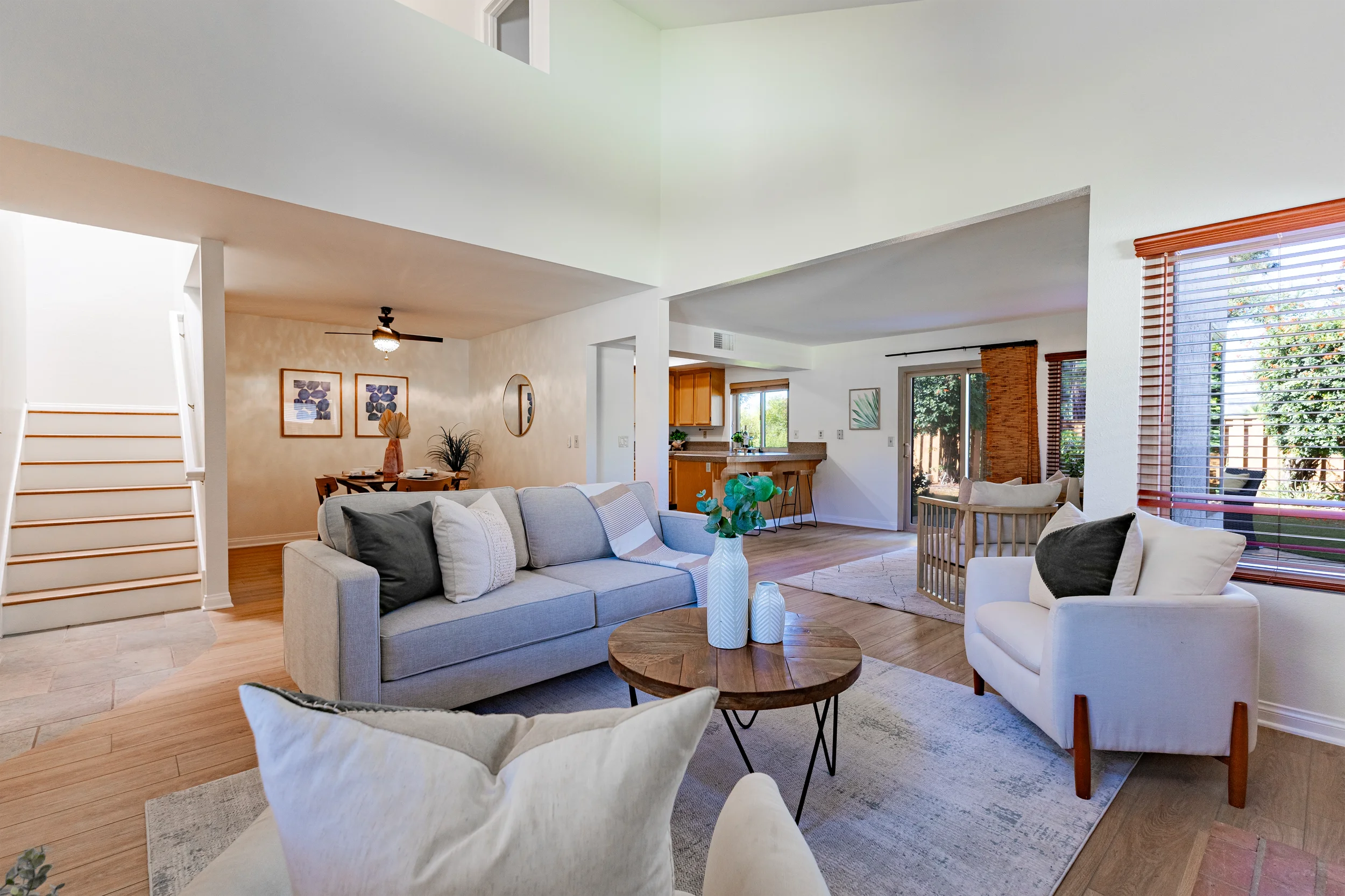 Bright living room with gray sofa, white armchair, wooden coffee table with vases, large windows with blinds, and open kitchen in background.
