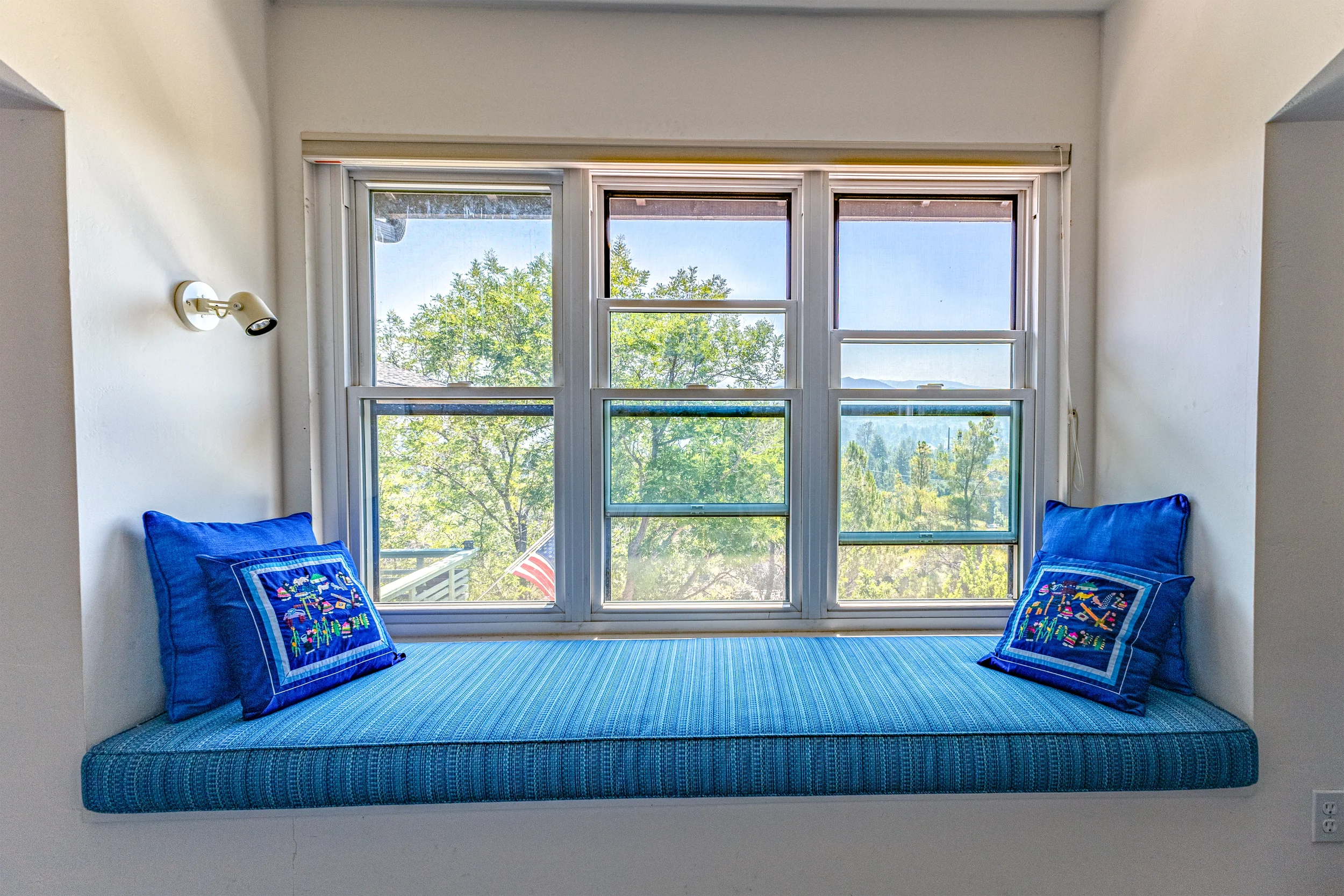 Sunlit window seat with blue cushion and two embroidered pillows, overlooking trees and distant hills.